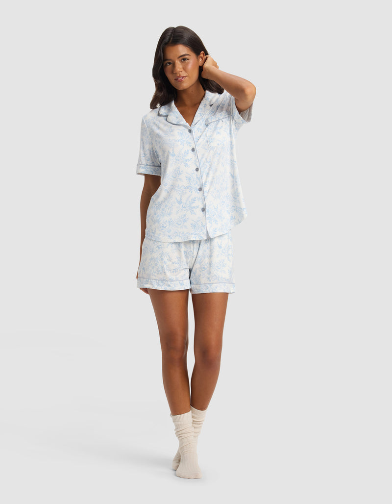 A woman stands in a relaxed pose wearing the Cozy Earth Women's Bamboo Stretch-Knit Short Sleeve Pajama Set with matching shorts and cream socks against a plain light background.