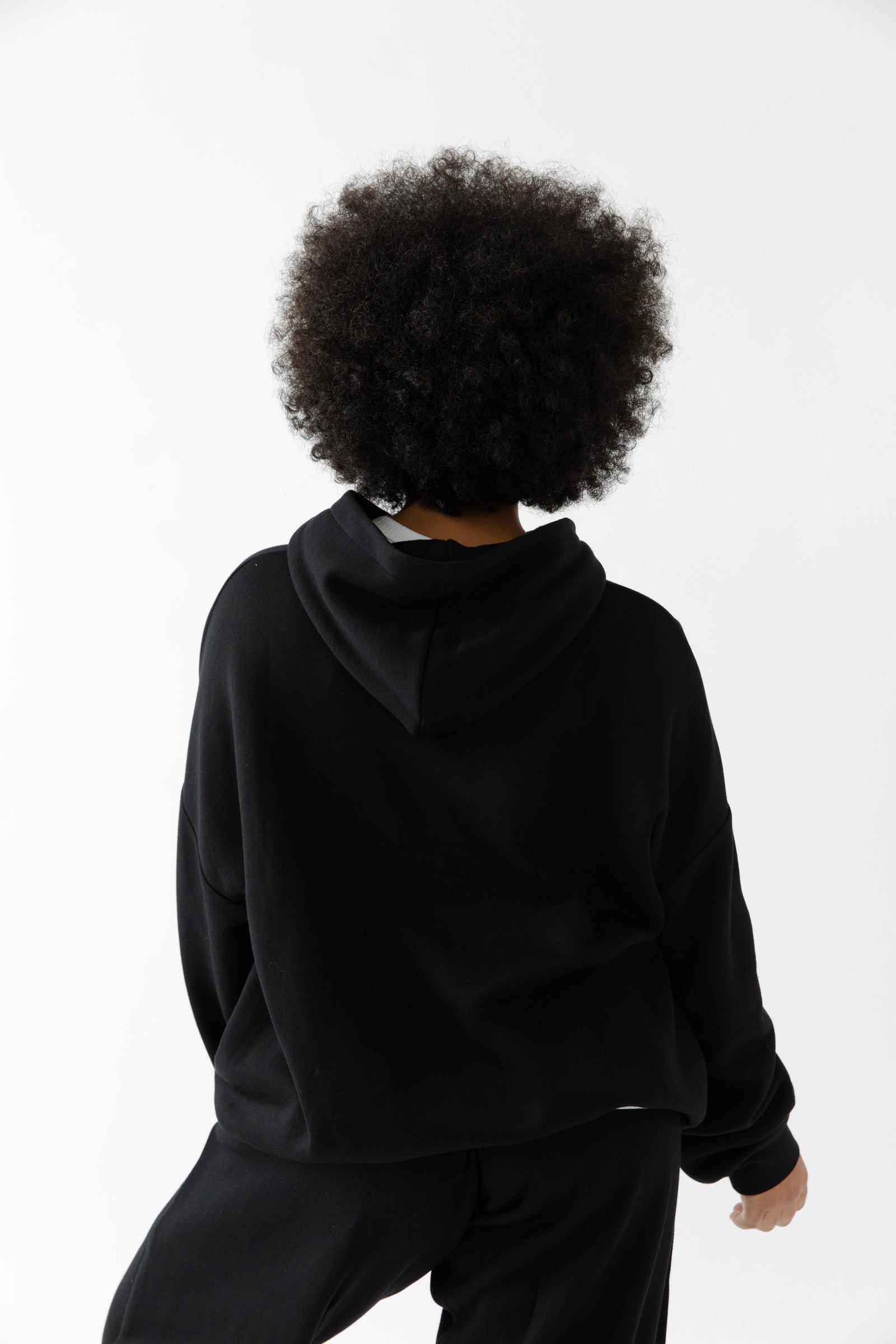 Model with curly hair wearing the Cozy Earth Women's CityScape Hoodie & Sweatpant Set in black, shown from the back against a plain white background. |Color:Black