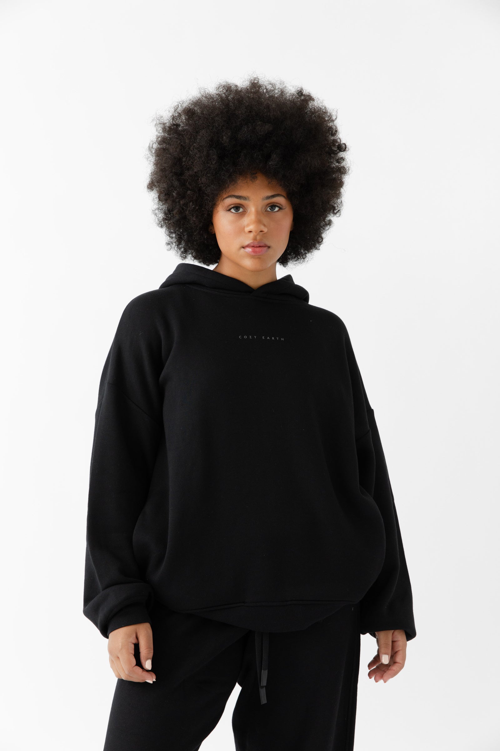 A woman with curly hair models the Cozy Earth Women's CityScape Hoodie & Sweatpant Set in black, standing against a plain white background and looking directly at the camera with a neutral expression. |Color:Black