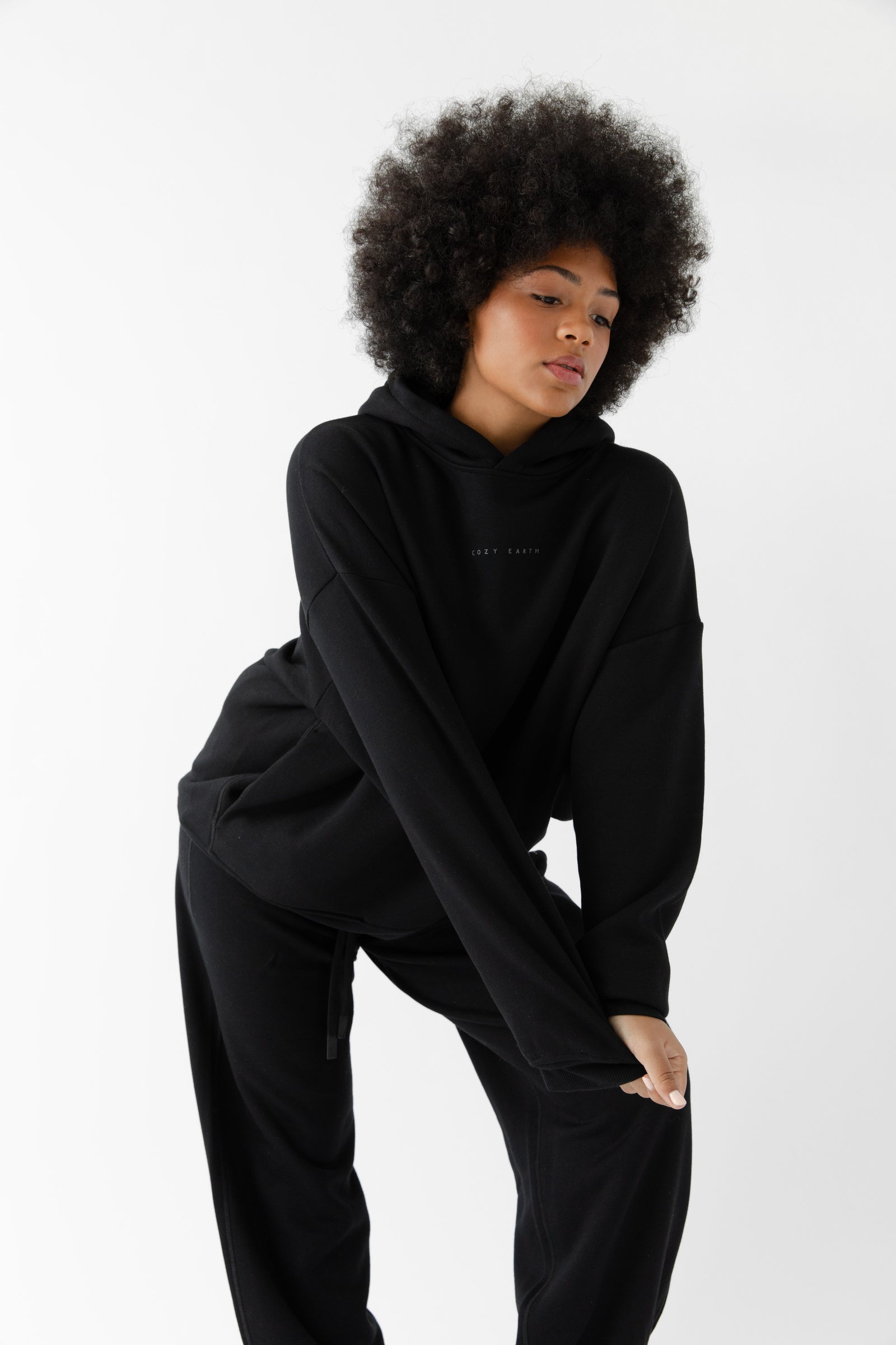 A woman with curly hair models the Cozy Earth Women's CityScape Hoodie & Sweatpant Set in black, leaning forward with one hand on her knee, posed against a plain white background. 