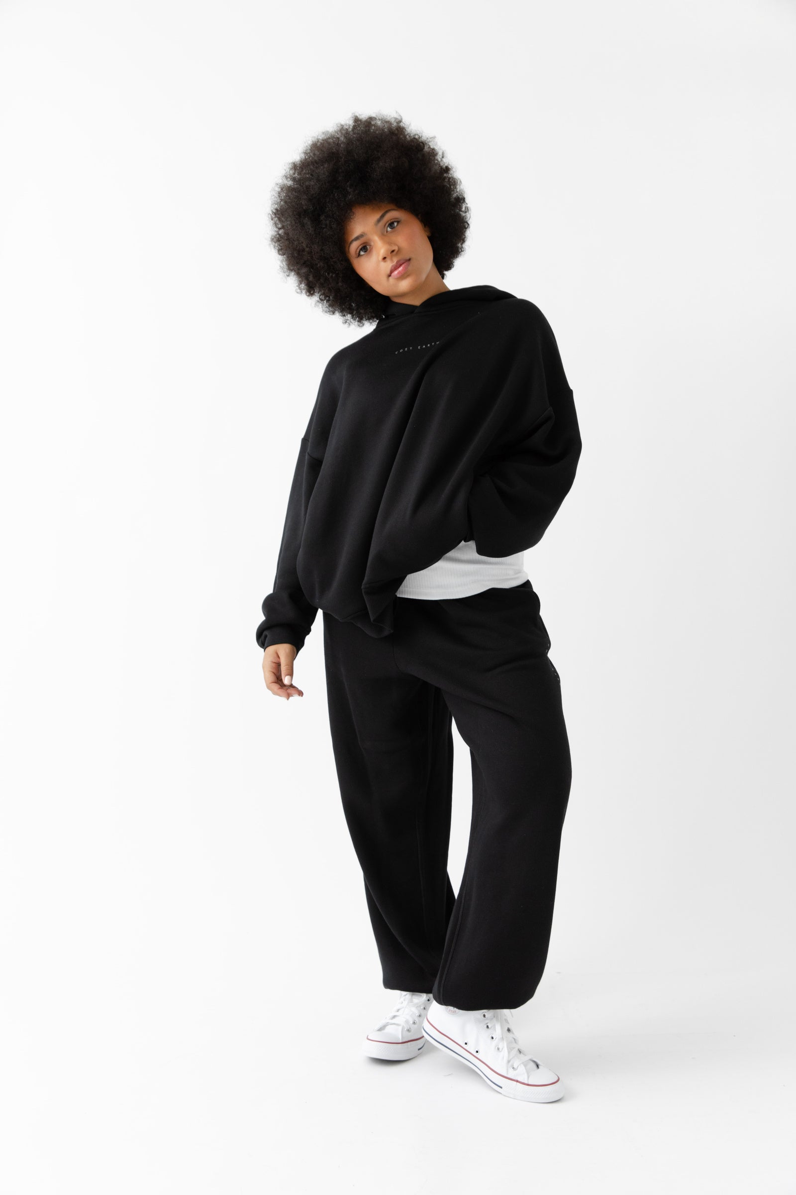 A woman with curly hair models the Cozy Earth Women's CityScape Hoodie & Sweatpant Set in black, paired with white sneakers, standing against a plain white background and gazing at the camera in a relaxed pose. |Color:Black