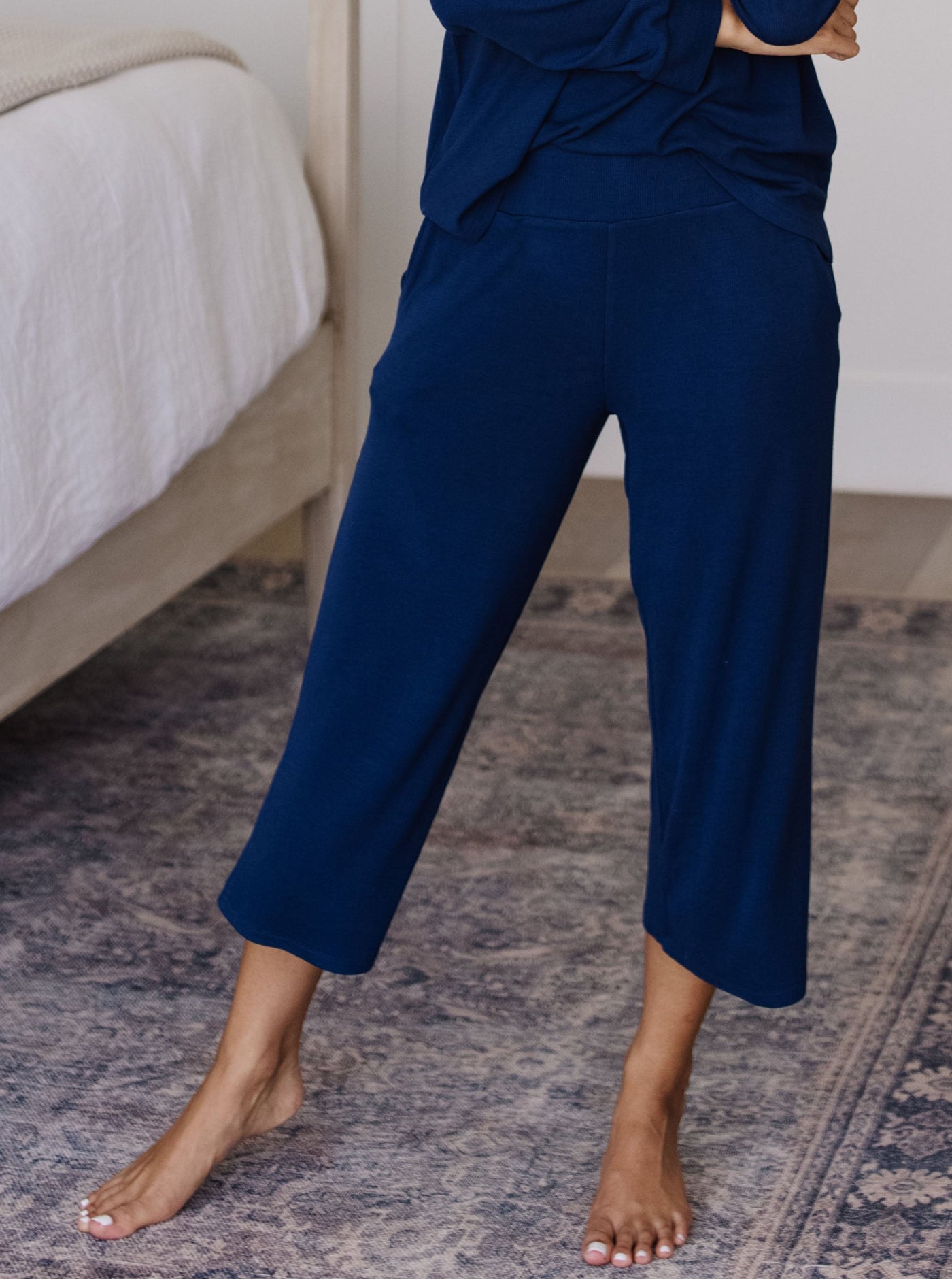 A woman stands barefoot on a patterned rug, wearing Cozy Earth’s Women’s Bamboo Rib-Knit Lounge Capri and matching top in navy blue, next to a bed with a white comforter. Her face is not visible.