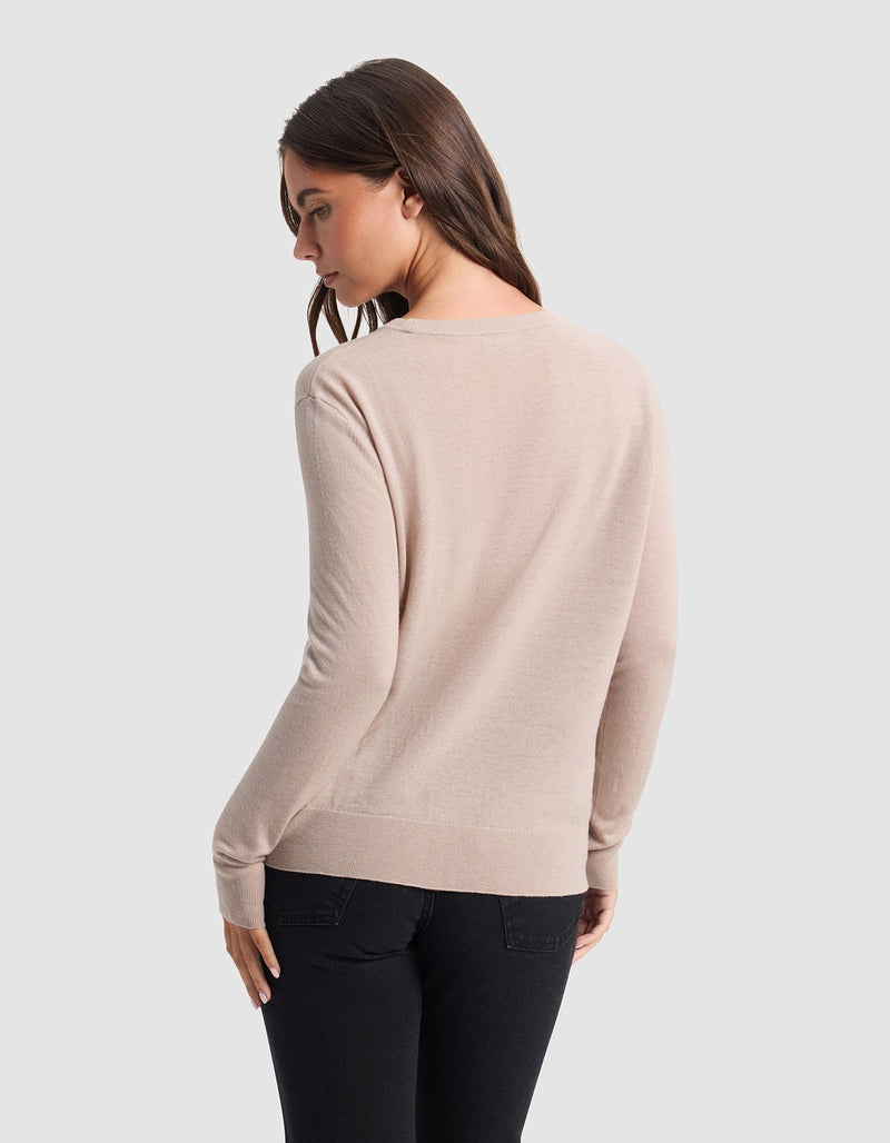 A woman with long brown hair wears the Cozy Earth Women's AirKnit Crewneck Sweater in light beige and black pants, standing with her back slightly turned and looking over her left shoulder against a plain light gray background.