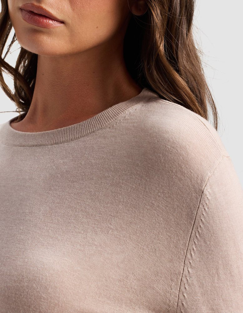 A close-up of a woman wearing the Cozy Earth Women's AirKnit Crewneck Sweater in light beige, showing her shoulder, part of her face, and wavy brown hair against a plain background.