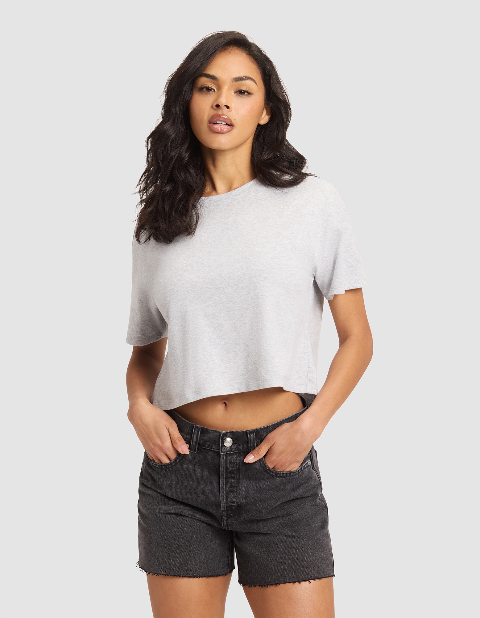 A woman with long dark hair models the Cozy Earth Women's All Day Cropped Tee in light gray and high-waisted black denim shorts. She stands against a plain light gray background, hands in pockets, looking at the camera. 