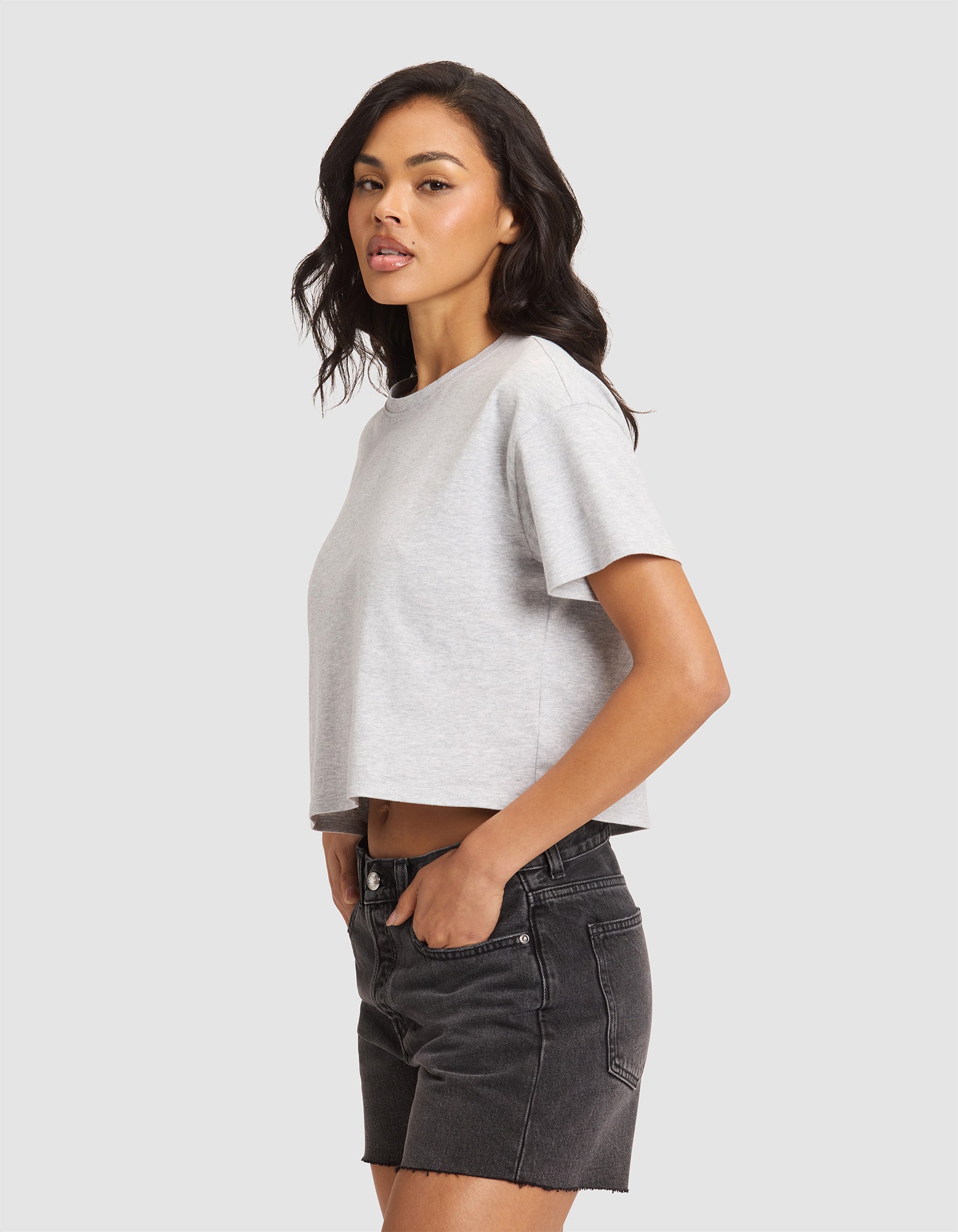 A woman with medium-length dark wavy hair wears the Cozy Earth Women's All Day Cropped Tee in light gray and black denim shorts, standing sideways against a plain light background with a relaxed, confident expression. 