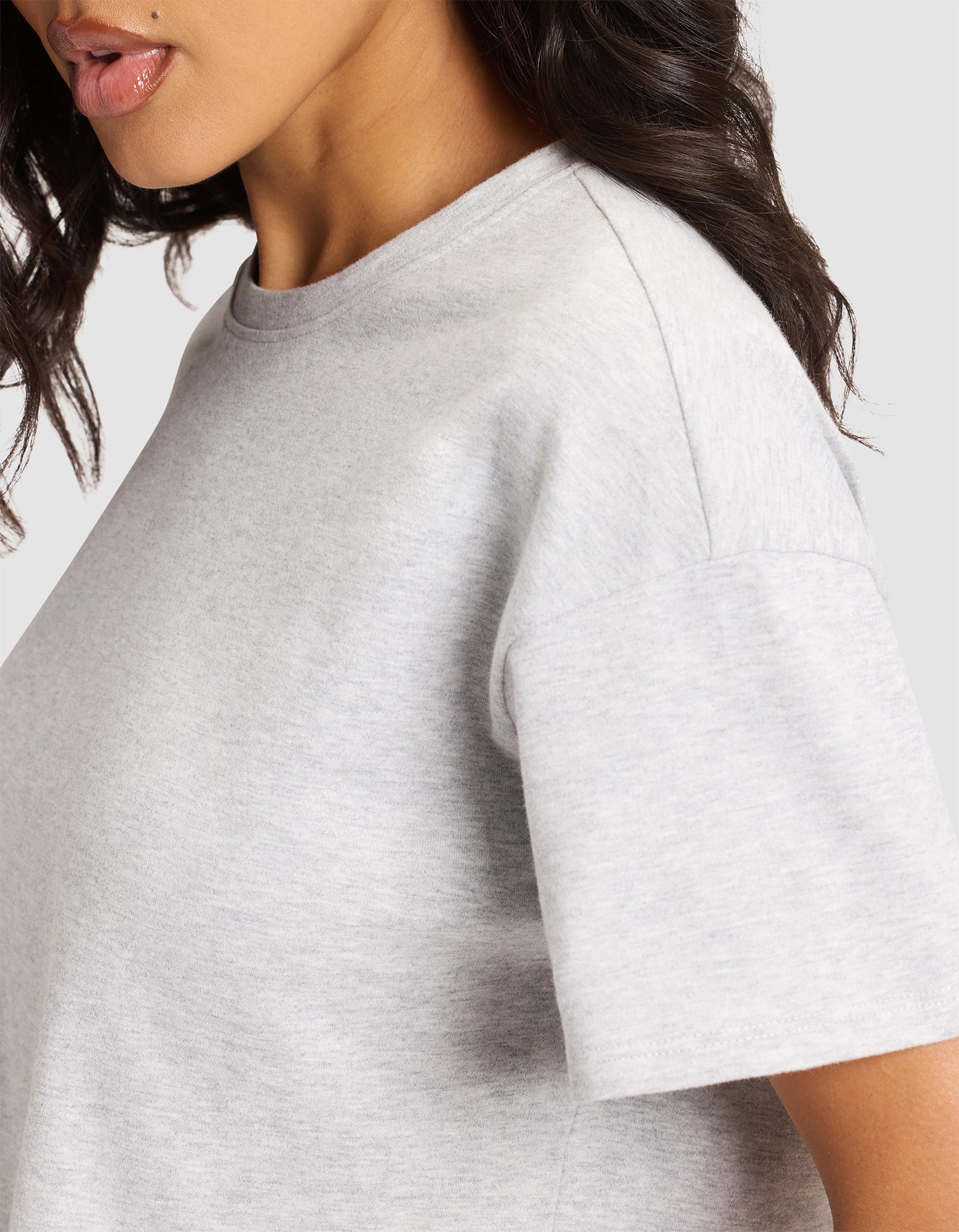 A close-up shows a woman in the Cozy Earth Women's All Day Cropped Tee in light grey, with only part of her face, neck, and upper torso visible as her wavy dark hair falls over her shoulder. 
