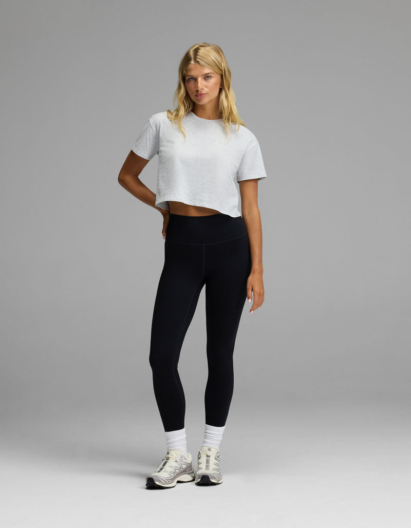 A woman stands against a plain gray background wearing the Cozy Earth Women’s All Day Cropped Tee in white, paired with black leggings, white socks, and sneakers. She has blonde hair and poses casually with one hand on her hip.