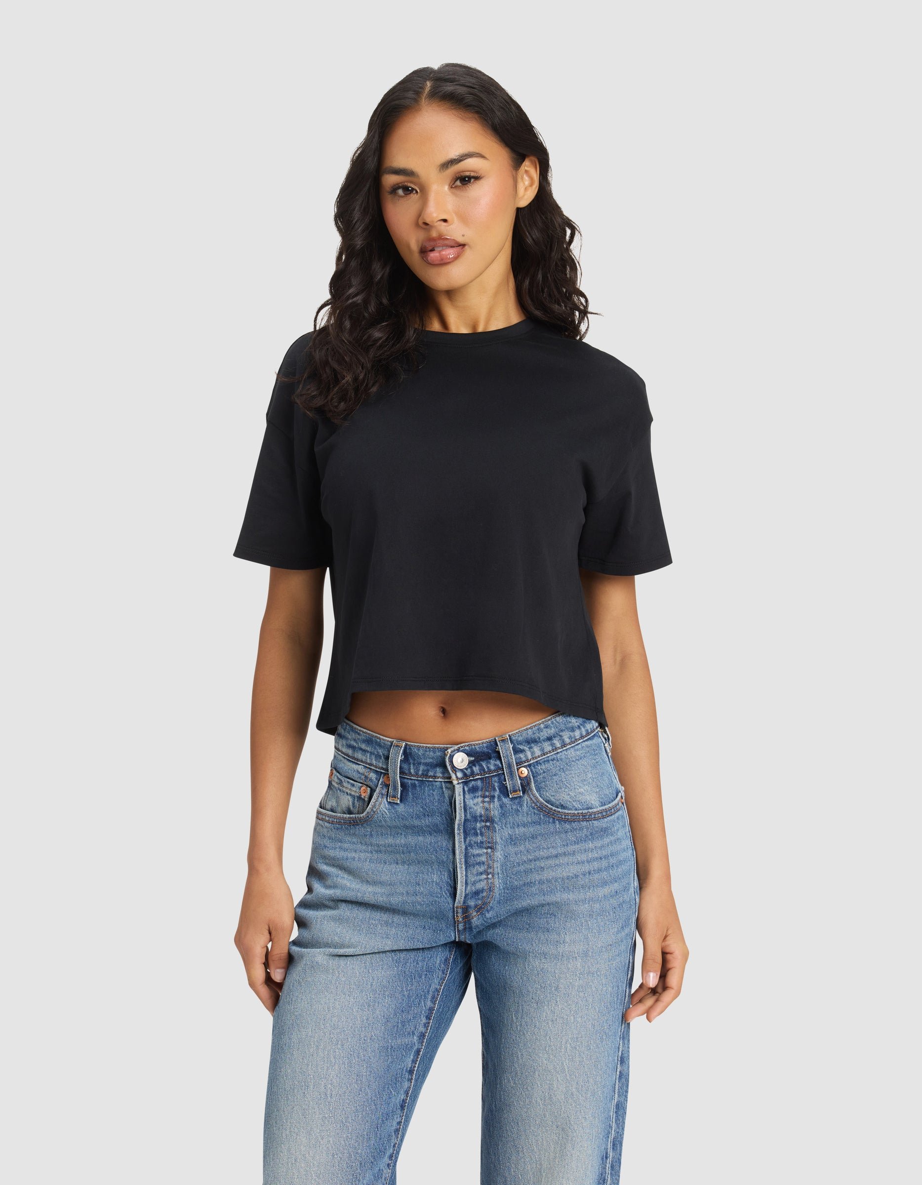 A woman with long, wavy hair wears the black Cozy Earth Women's All Day Cropped Tee and blue jeans, standing against a plain light gray background. |Color:Jet Black