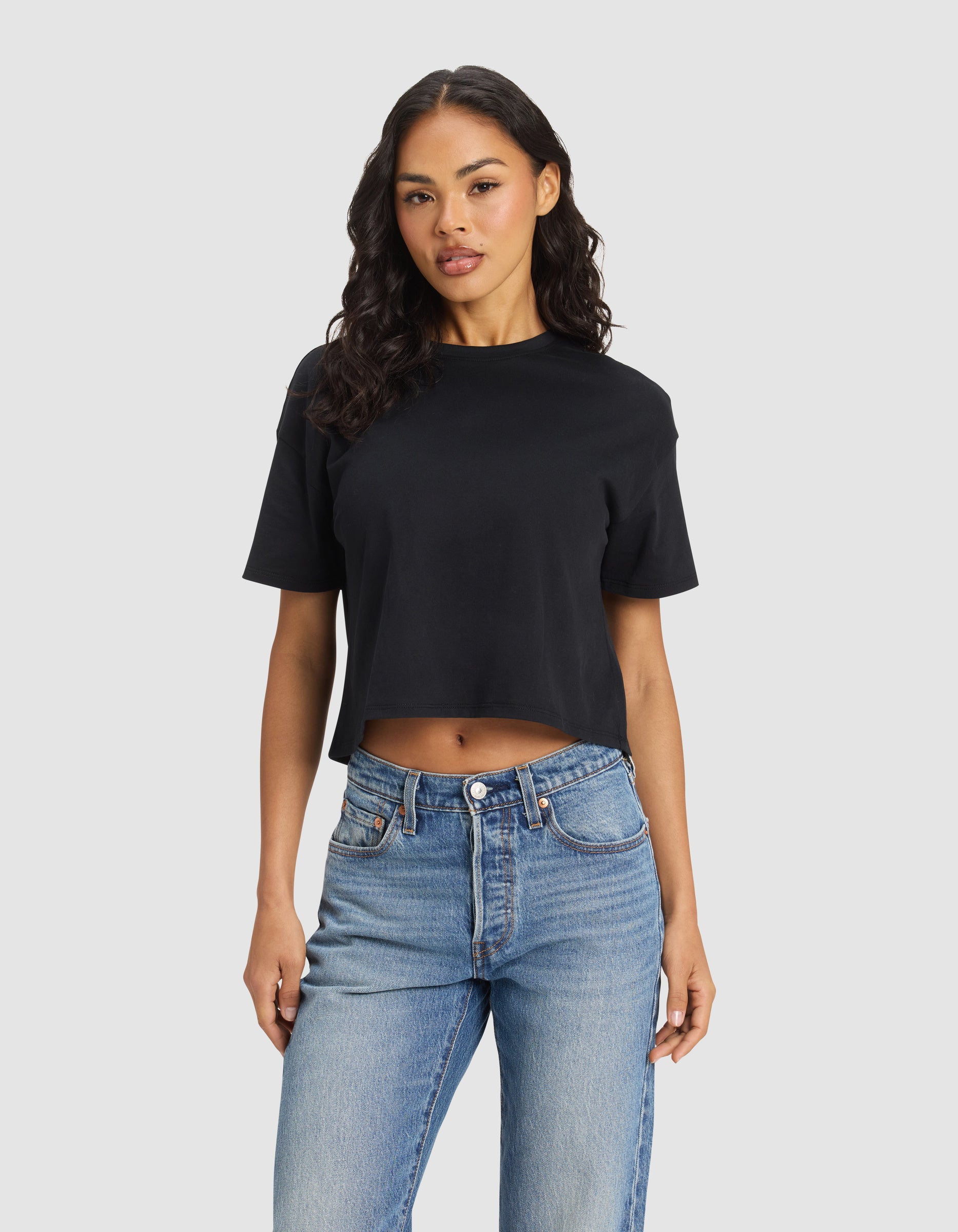 A woman with long, wavy hair wears the black Cozy Earth Women's All Day Cropped Tee and blue jeans, standing against a plain light gray background. |Color:Jet Black