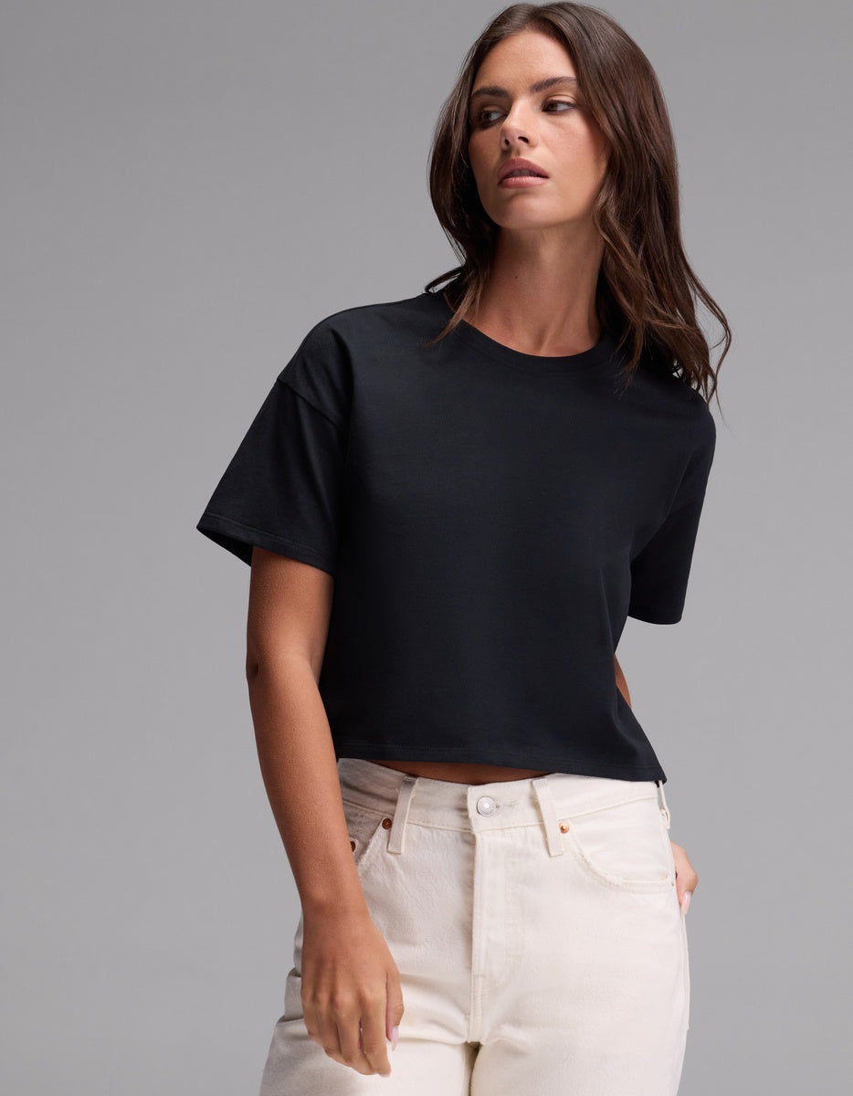 A woman stands against a plain gray background, wearing the Cozy Earth Women’s All Day Cropped Tee in Jet Black paired with high-waisted white jeans, with one hand casually placed in her pocket.