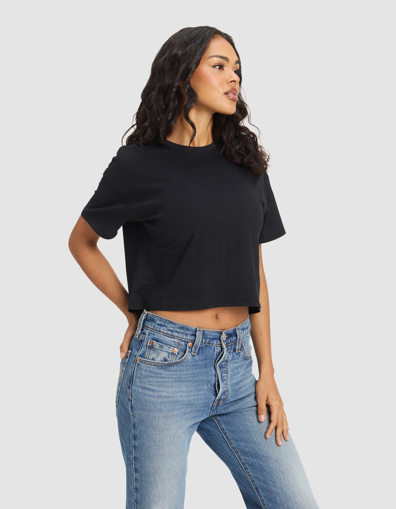 A woman with wavy dark hair wears the Cozy Earth Women's All Day Cropped Tee in black and high-waisted blue jeans, standing against a light background and looking to the side with one hand on her hip.