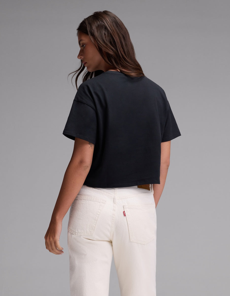 A woman with long brown hair stands facing away against a plain gray background, wearing the Cozy Earth Women’s All Day Cropped Tee in black paired with white jeans.