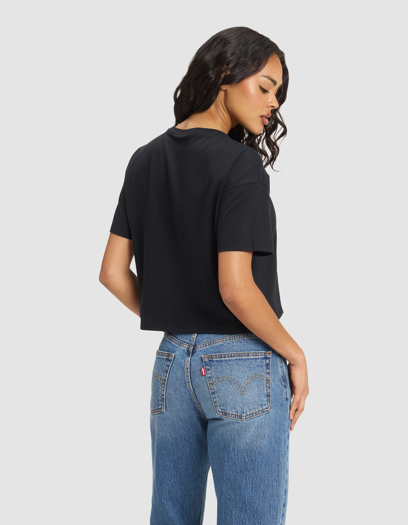 A woman with long wavy hair models the Cozy Earth Women's All Day Cropped Tee in black and blue jeans, turned slightly to the side to display the back of the shirt, with one hand on her hip.
