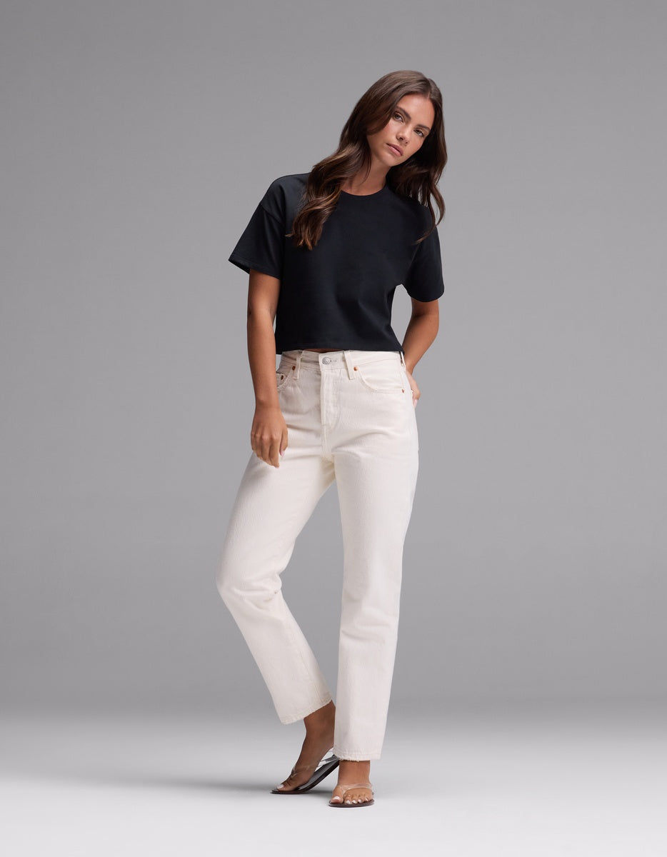 A woman with long brown hair wears the Cozy Earth Women’s All Day Cropped Tee in black, white high-waisted jeans, and brown sandals. She stands against a plain gray background with her hands in her pockets.