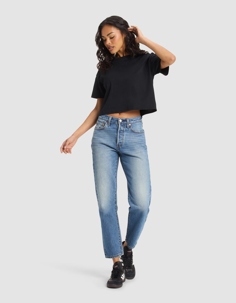 A woman with long curly hair wears the Cozy Earth Women's All Day Cropped Tee in black, paired with light blue high-waisted jeans and black sneakers, standing against a plain light background.