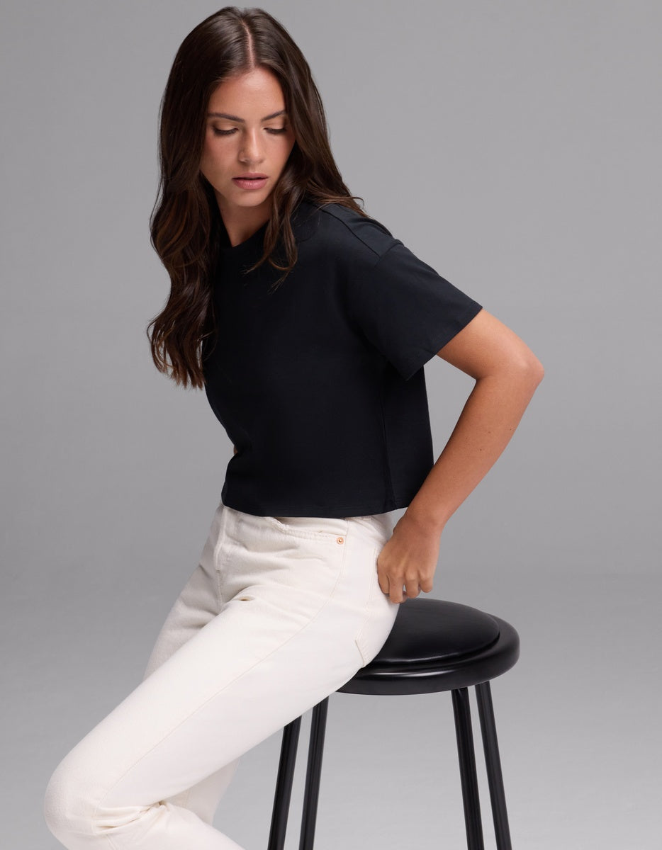 A woman with long brown hair sits sideways on a black stool against a plain gray background, looking down to her left. She is wearing the Cozy Earth Women’s All Day Cropped Tee in black and white pants.