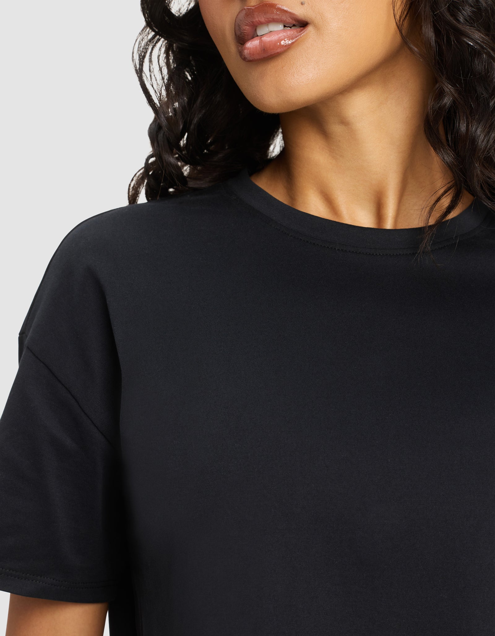 A close-up shows a woman with wavy dark hair wearing the Cozy Earth Women's All Day Cropped Tee in black; her lips, neck, and shoulder are visible against a light background.