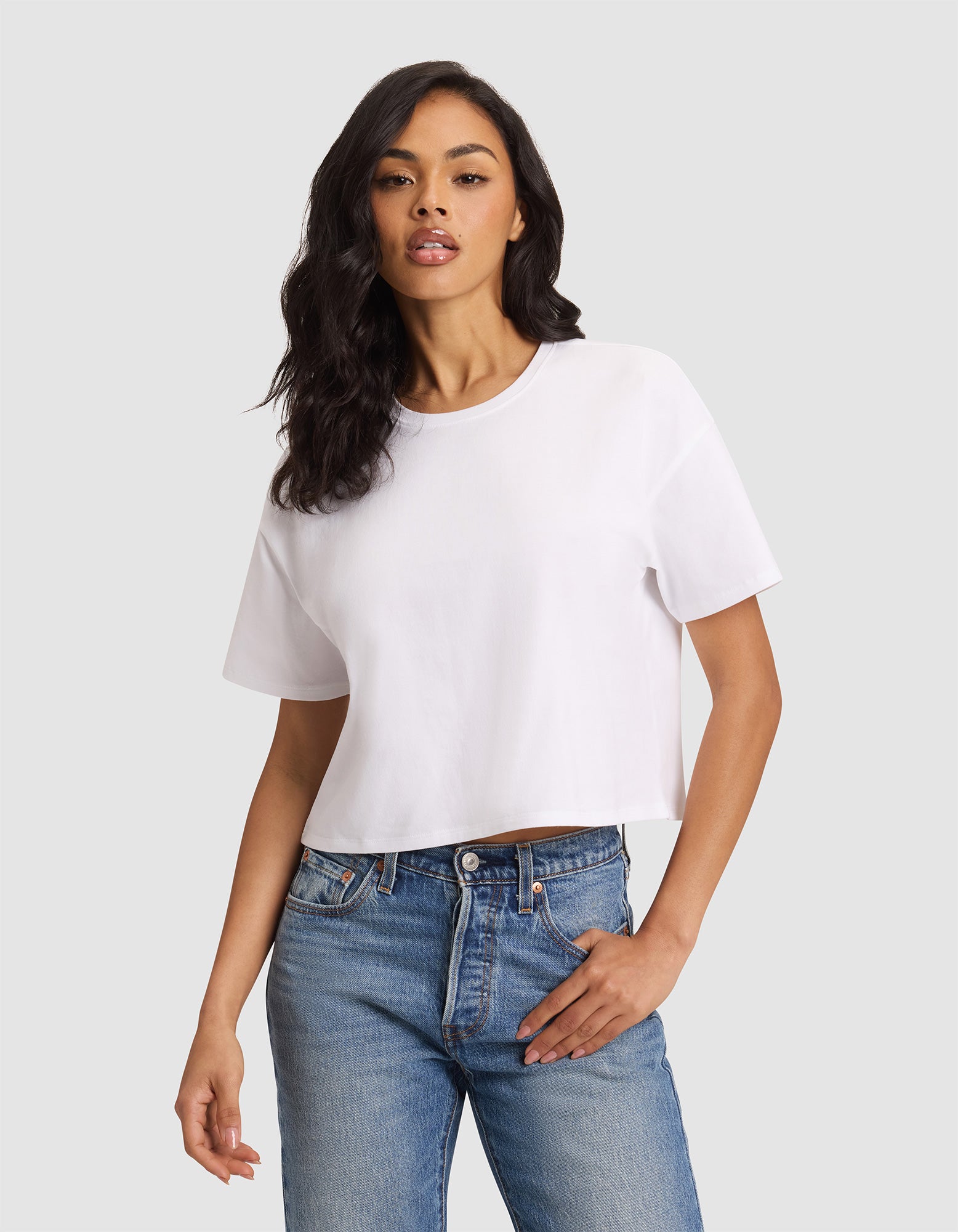 A woman with long dark hair models the Cozy Earth Women's All Day Cropped Tee in white with blue jeans, standing against a light gray background and posing with one hand in her pocket. 