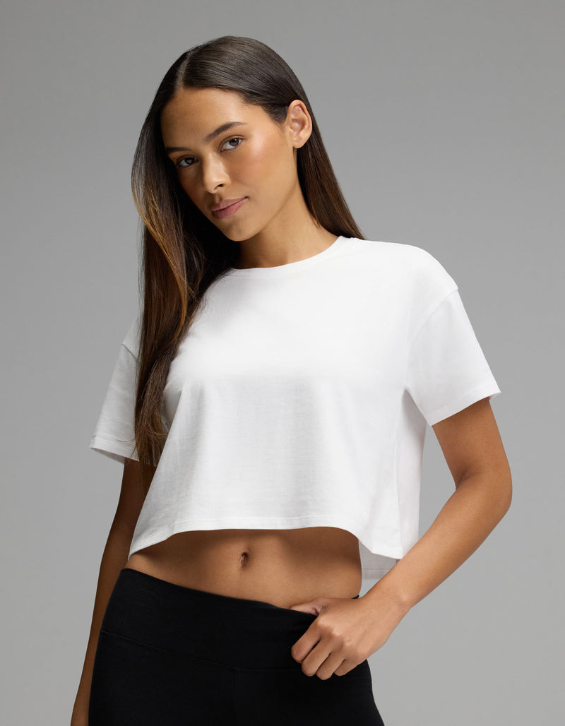 A woman with long brown hair stands confidently against a plain gray background, wearing the Cozy Earth Women’s All Day Cropped Tee in white paired with black pants.
