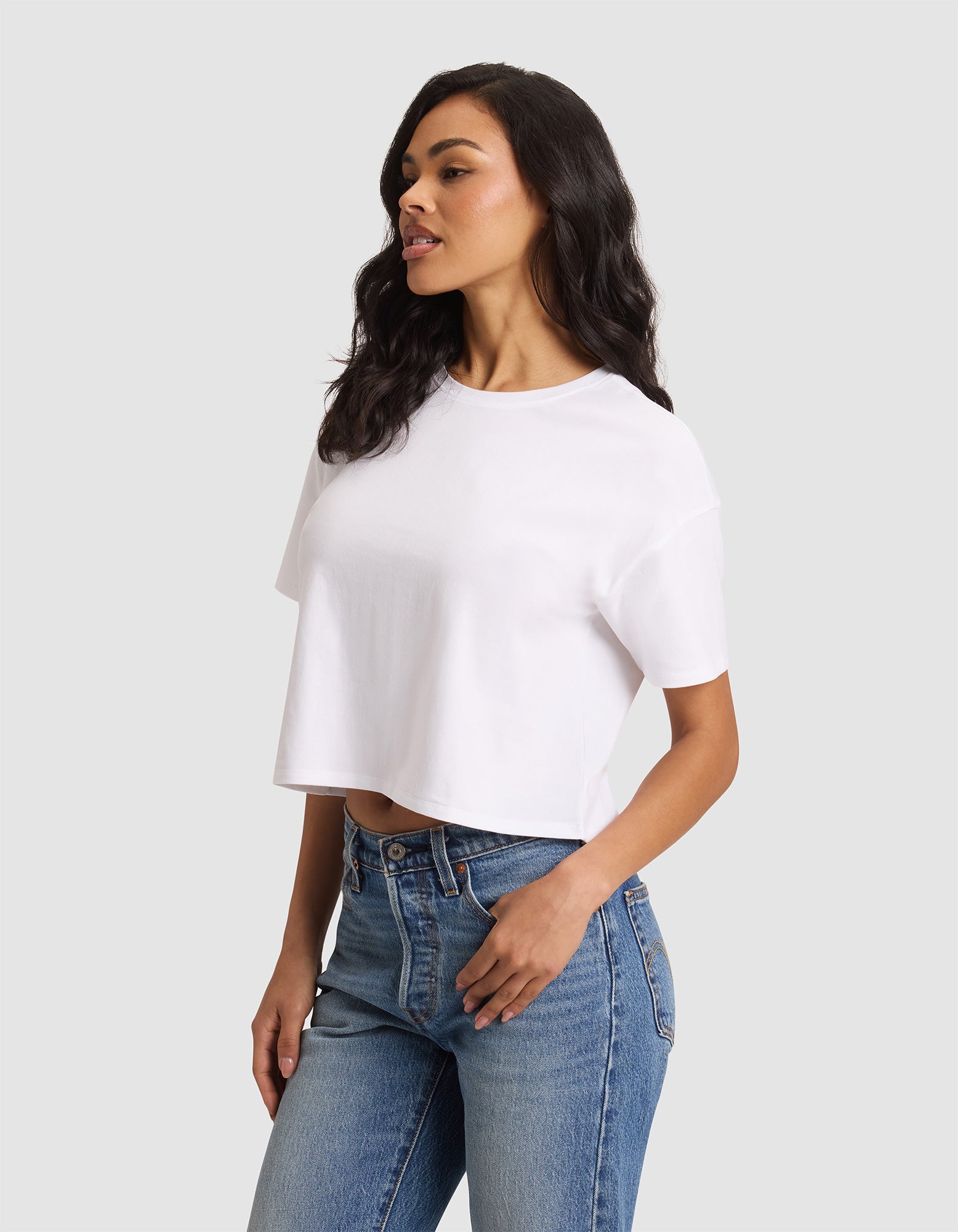 A woman wears the Cozy Earth Women's All Day Cropped Tee in white with blue jeans, standing against a light gray background and looking to the side with one hand on her hip. 