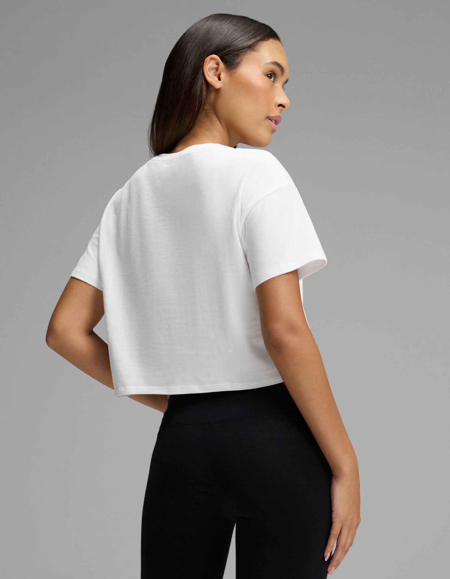 A woman with long dark hair models the Cozy Earth Women’s All Day Cropped Tee in white, paired with black leggings. She stands sideways, looking over her shoulder against a plain gray background.|Color:White