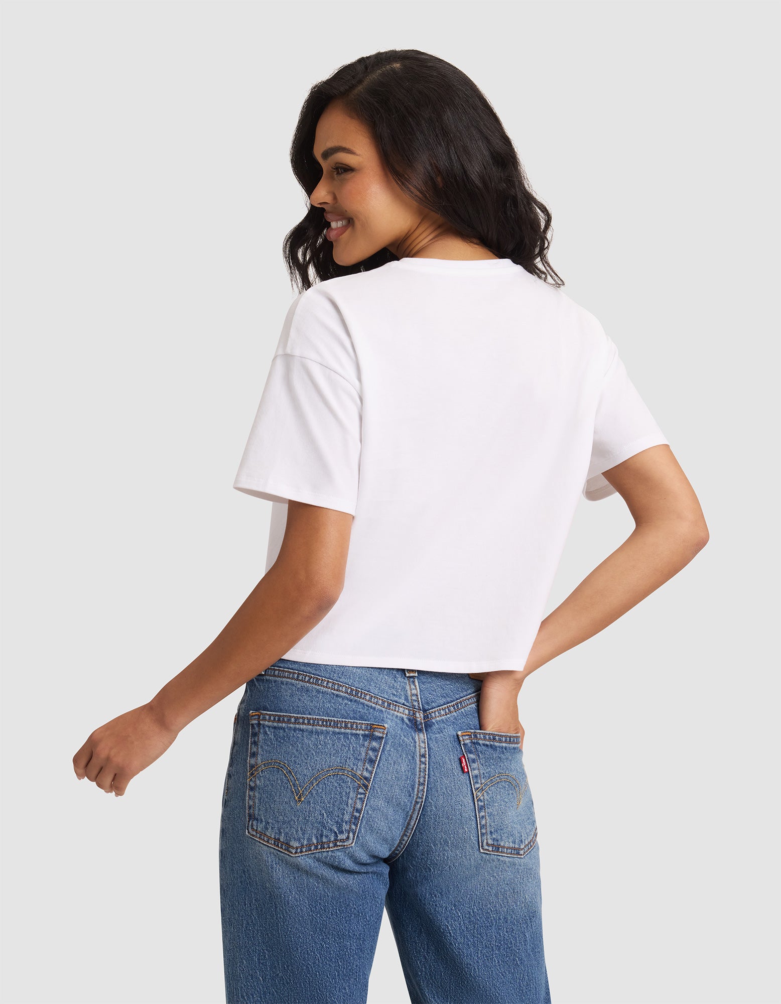 A woman with long dark hair wears the Cozy Earth Women's All Day Cropped Tee in white and blue jeans, standing sideways with a smile and one hand in her back pocket against a light gray background. 