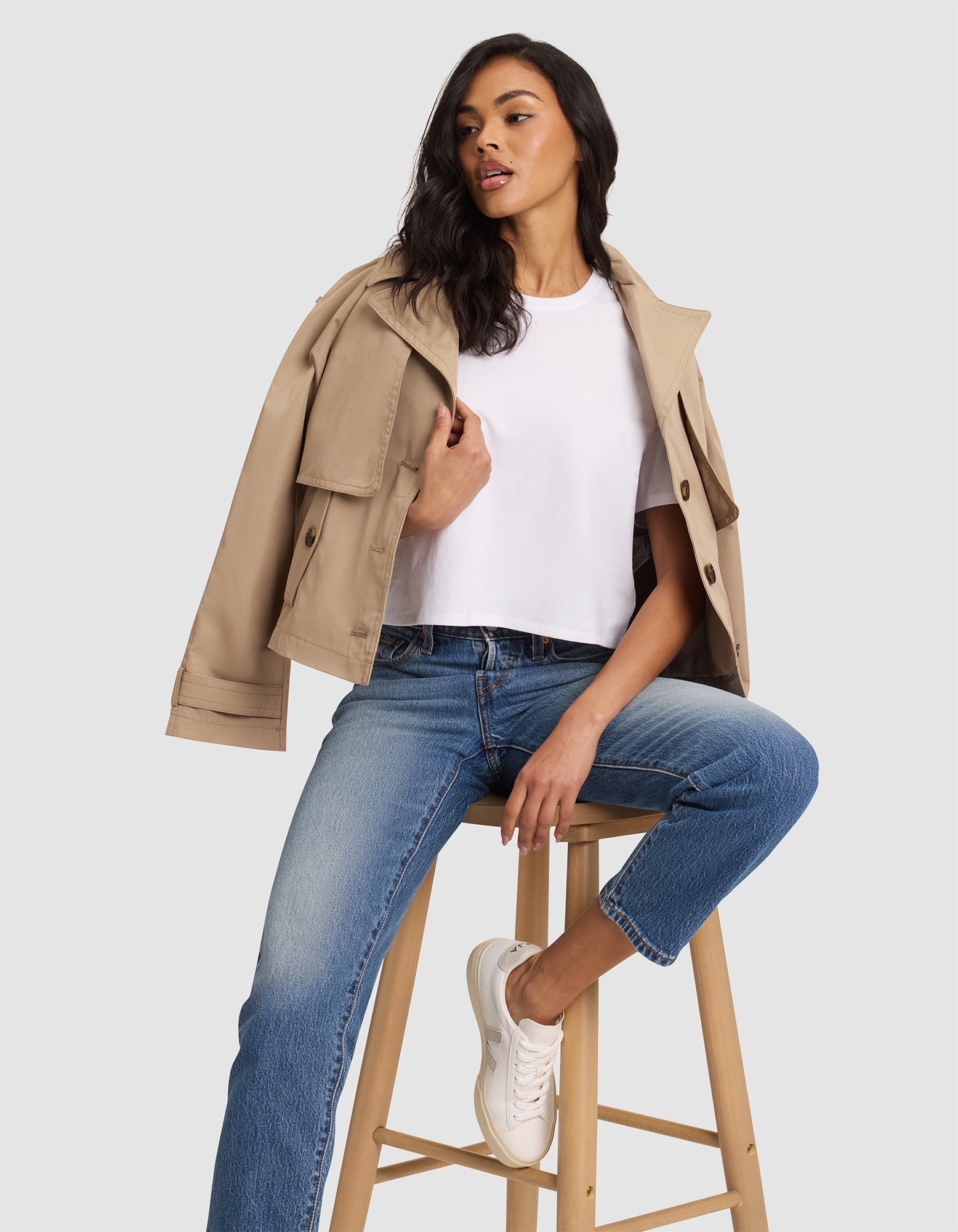 A woman poses confidently on a wooden stool against a light gray background, wearing the Cozy Earth Women's All Day Cropped Tee, paired with a beige trench coat, blue jeans, and white sneakers. 