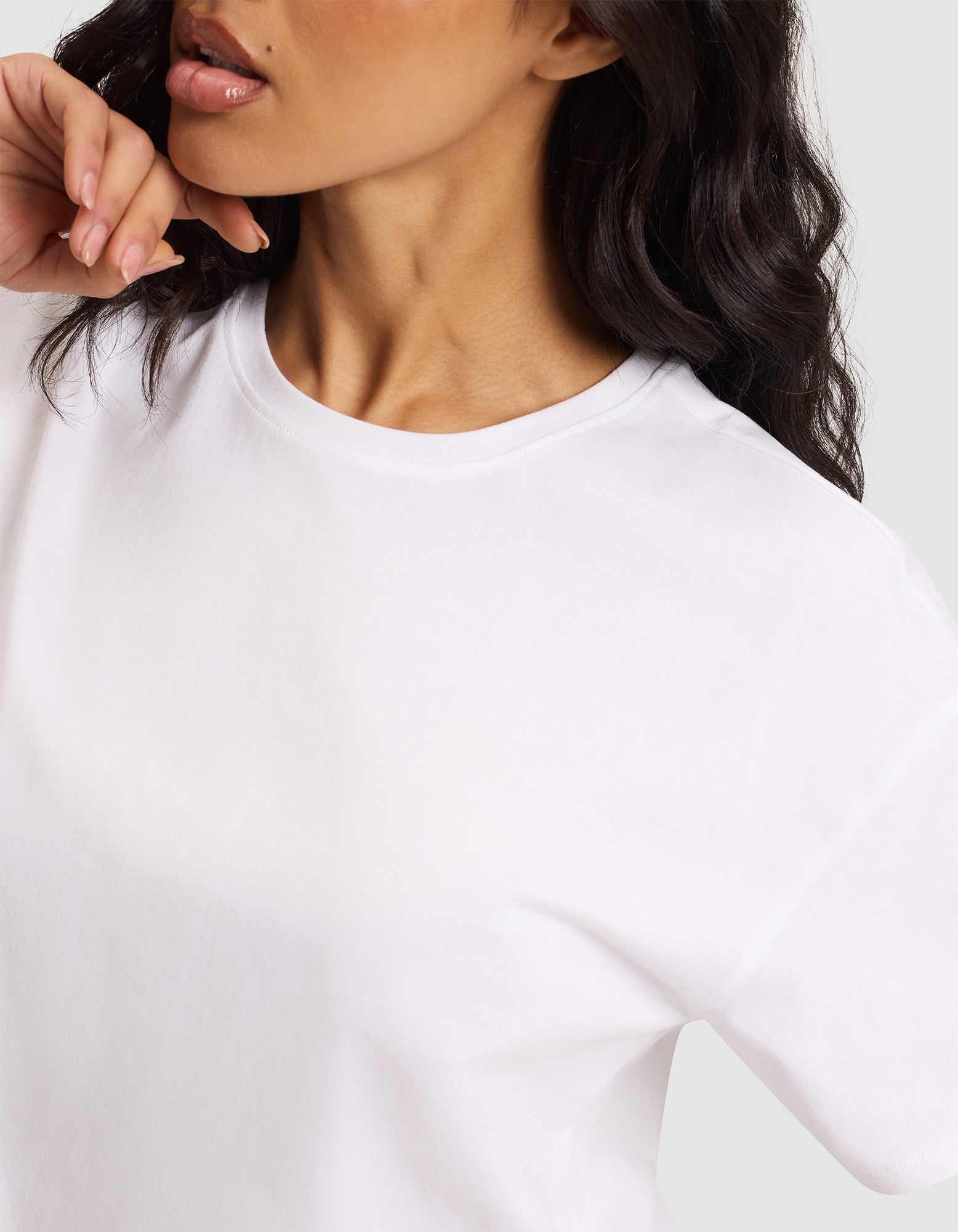A woman with wavy dark hair wears the Cozy Earth Women's All Day Cropped Tee in white. Her face is partially visible as she gently touches her chin with her left hand, set against a neutral background. 