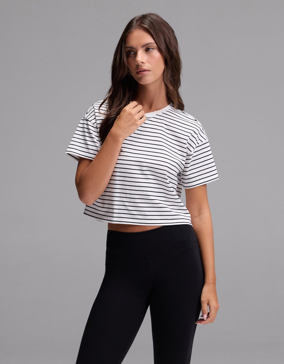 A woman with long brown hair is wearing the Cozy Earth Women’s All Day Cropped Tee in Jet Black Stripe and black leggings, standing against a plain gray background.