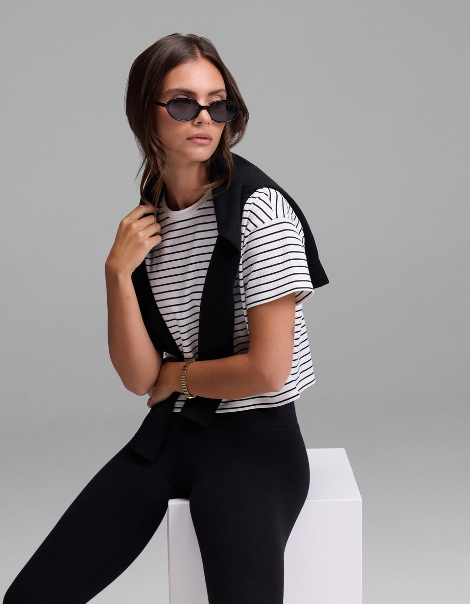 A woman wearing black sunglasses, black pants, and the Cozy Earth Women’s All Day Cropped Tee sits on a white block with a black sweater draped over her shoulders against a plain light gray background.