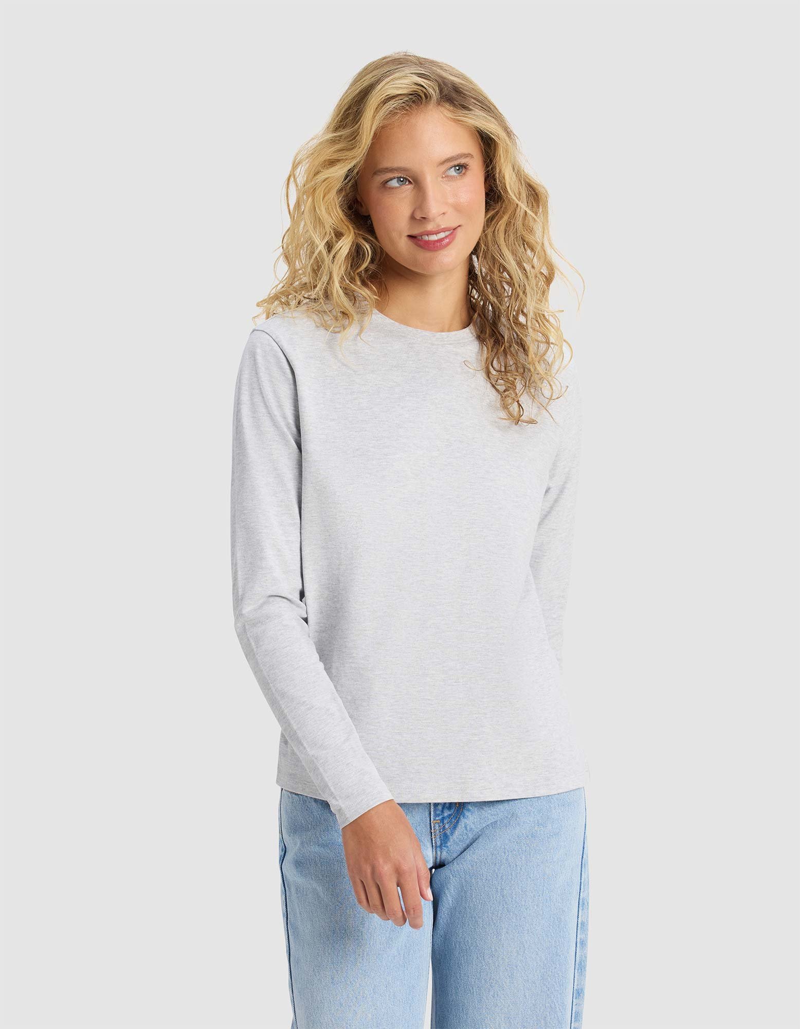 A woman with long curly blonde hair wears the Cozy Earth Women's All Day Long Sleeve Tee in light gray and light blue jeans, standing against a plain light background and smiling slightly.|Color:Heather Grey