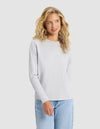 A woman with long curly blonde hair wears the Cozy Earth Women's All Day Long Sleeve Tee in light gray and light blue jeans, standing against a plain light background and smiling slightly.|Color:Heather Grey