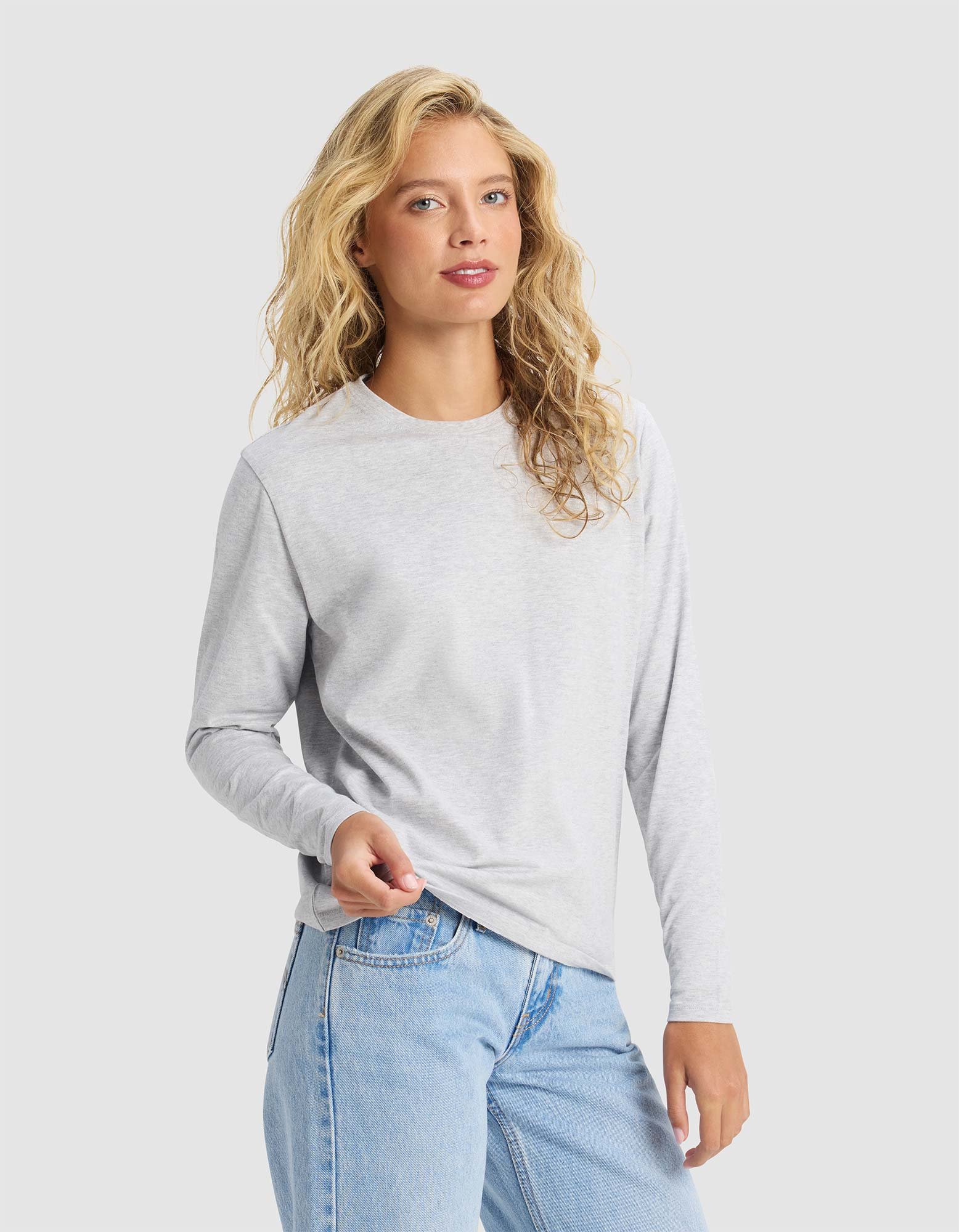 A woman with long, wavy blonde hair models the Cozy Earth Women's All Day Long Sleeve Tee in light gray and light blue jeans, standing against a plain light background and holding the shirt's hem.