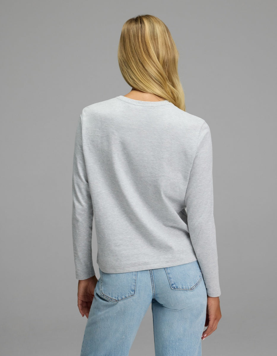 A woman with long blonde hair stands facing away, wearing the Cozy Earth Women’s All Day Long Sleeve Tee in light gray and light blue jeans against a plain gray background.|Color:Heather Grey