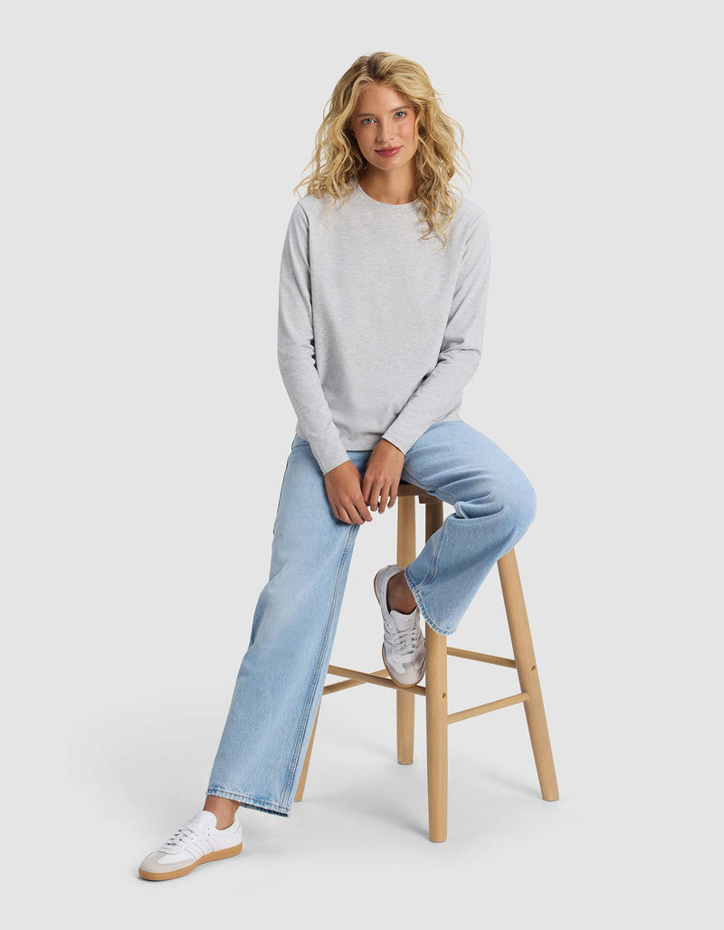 A woman with long curly blonde hair sits casually on a wooden stool against a plain light background, wearing Cozy Earth's Women's All Day Long Sleeve Tee in light gray, paired with light blue jeans and white sneakers.