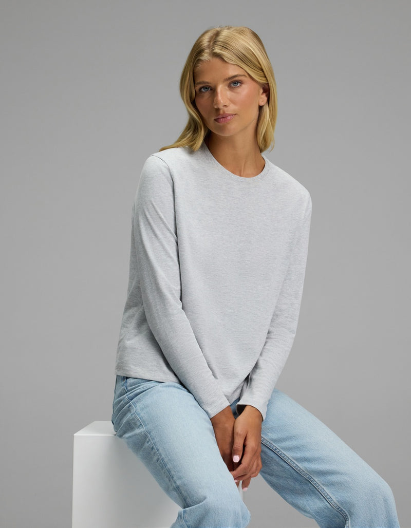 A woman with blonde hair sits on a white block against a plain gray background, wearing the Cozy Earth Women’s All Day Long Sleeve Tee in light gray and light blue jeans.