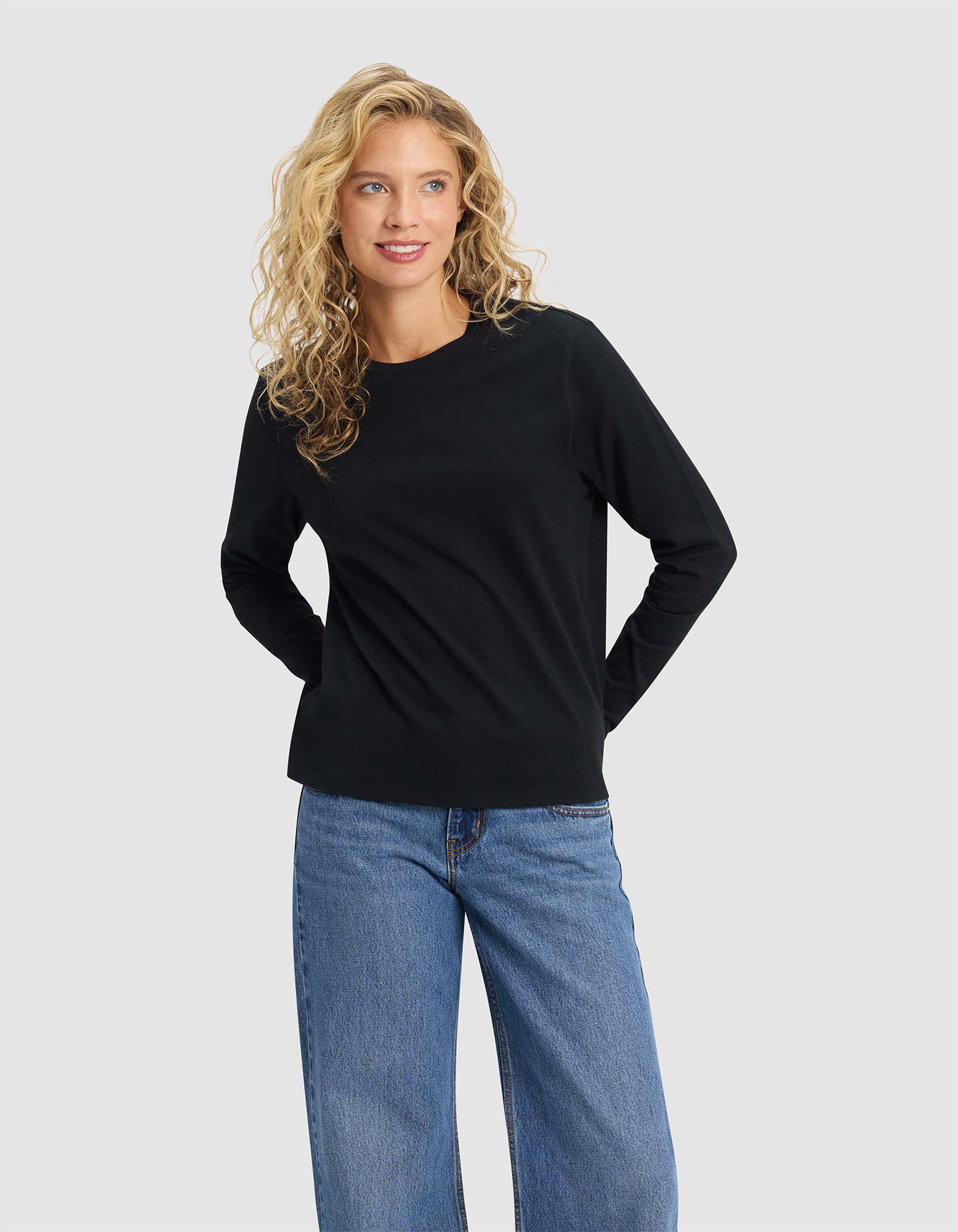 A woman with long curly blonde hair models the Cozy Earth Women's All Day Long Sleeve Tee in black, paired with blue jeans. She stands against a plain light grey background, smiling as she looks slightly to the side.
