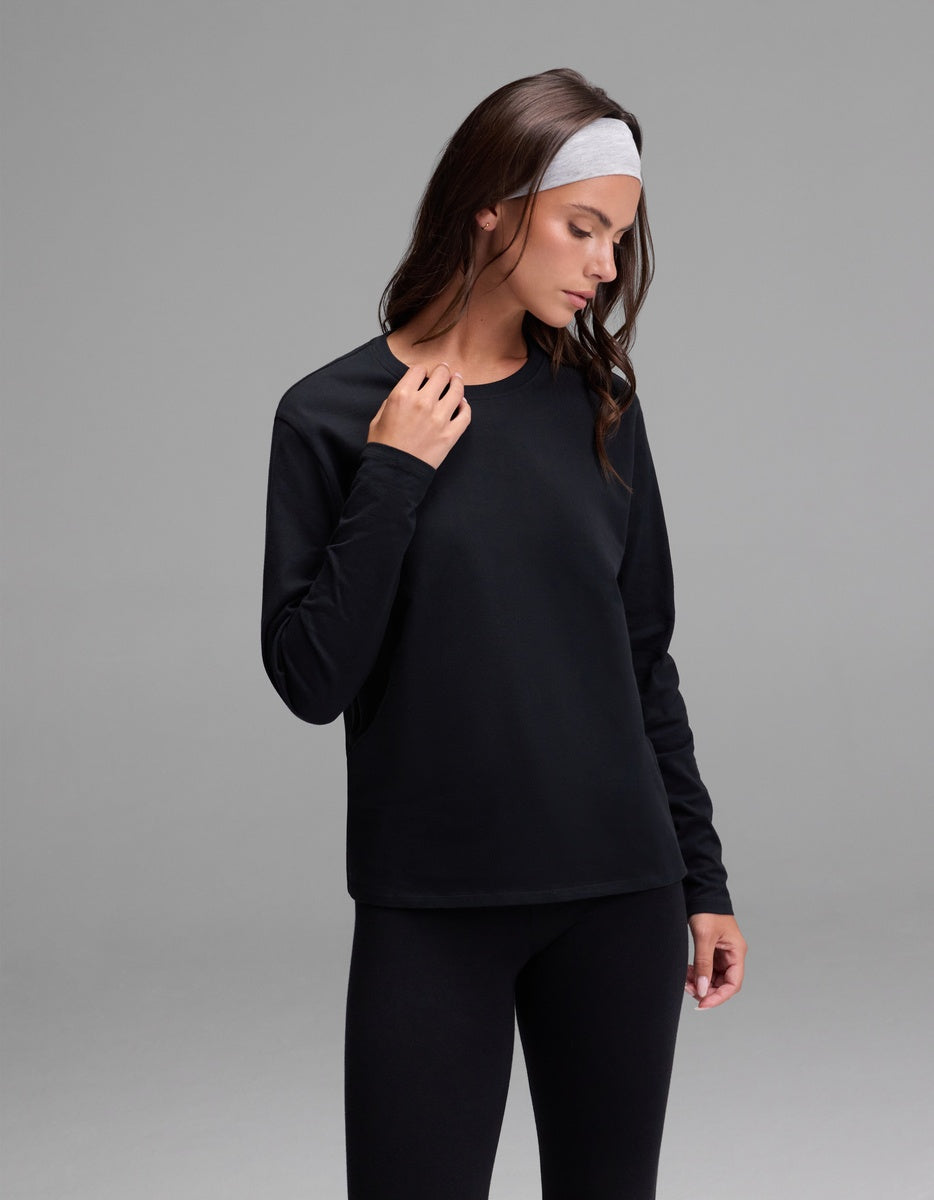 A woman wearing the Cozy Earth Women’s All Day Long Sleeve Tee in black, paired with black pants and a light gray headband, stands against a plain gray background, looking down and touching her shirt near her neck.|Color:Jet Black
