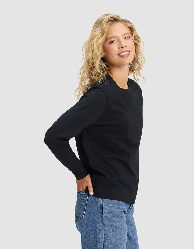 A woman with wavy blonde hair models the Cozy Earth Women's All Day Long Sleeve Tee in black with blue jeans, smiling sideways at the camera against a plain light gray background.