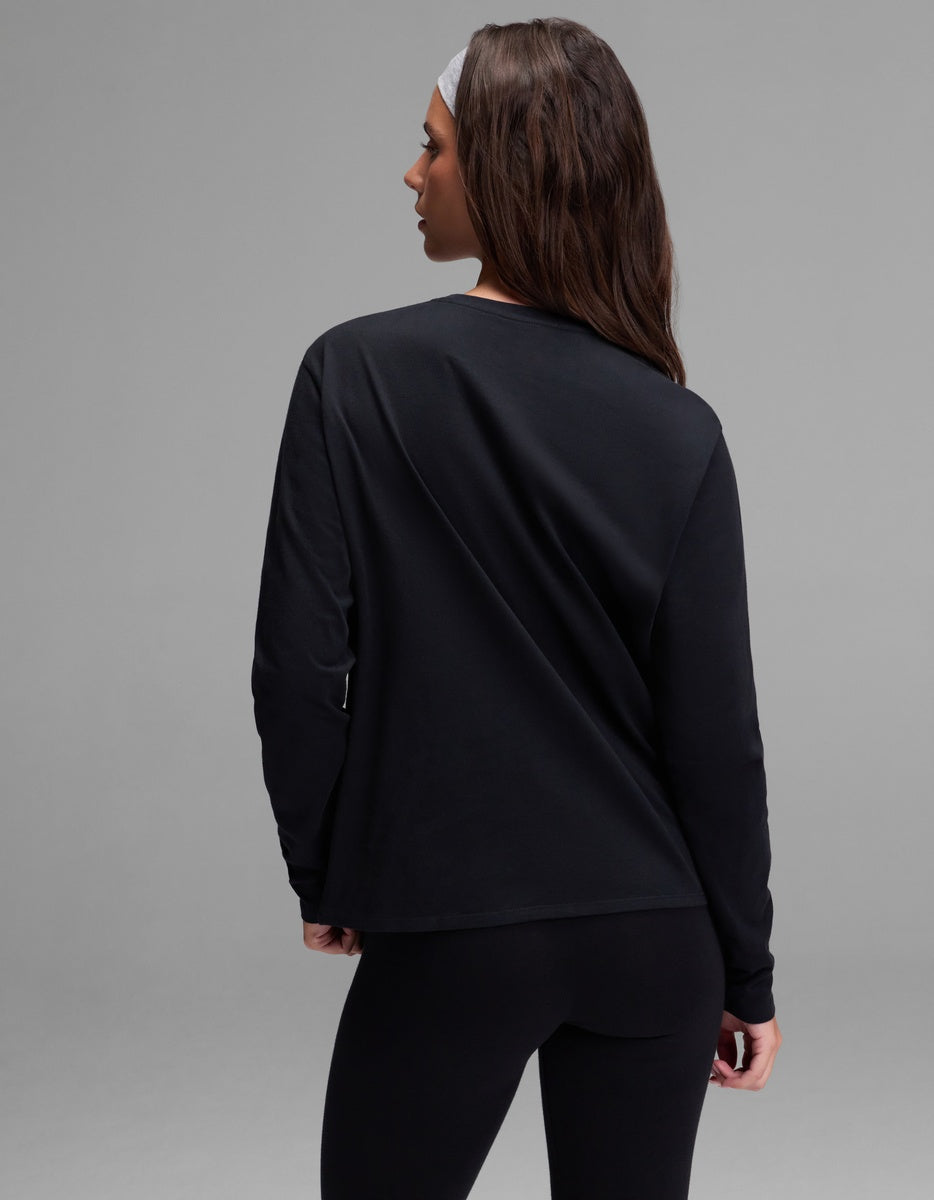 A woman with long brown hair, wearing Cozy Earth’s Women’s All Day Long Sleeve Tee in black and matching black leggings, stands facing away and looks over her left shoulder. She has a white headband and is set against a plain gray background.
