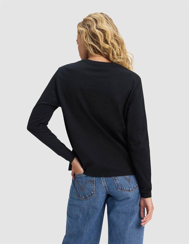 A woman with long, wavy blond hair is seen from the back wearing Cozy Earth’s Women's All Day Long Sleeve Tee in black with blue jeans. She poses against a plain light gray background, one hand on her hip and the other at her side.