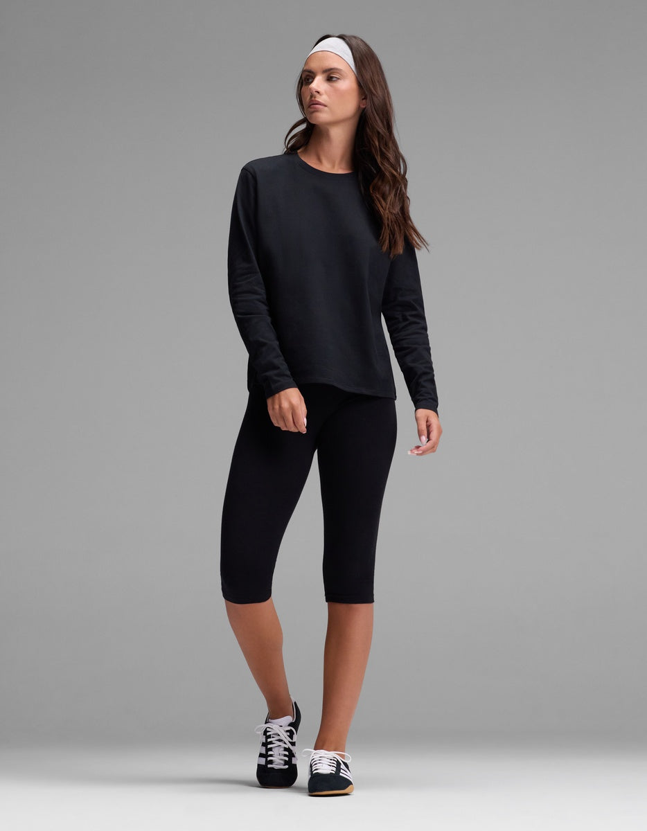 A woman wearing the Cozy Earth Women’s All Day Long Sleeve Tee in black, paired with black knee-length leggings, sneakers, and a grey headband, stands against a plain gray background, looking slightly to the side.