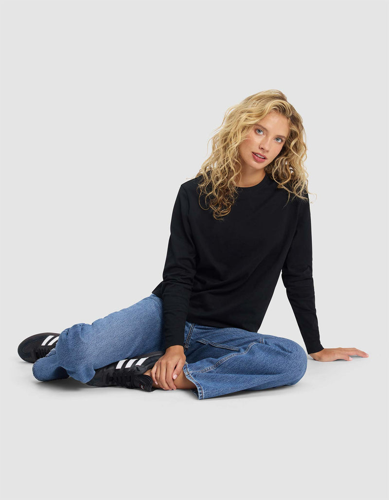 A woman with long curly blonde hair, wearing Cozy Earth’s Women's All Day Long Sleeve Tee in black, blue jeans, and black sneakers, sits casually on the floor against a plain light gray background.