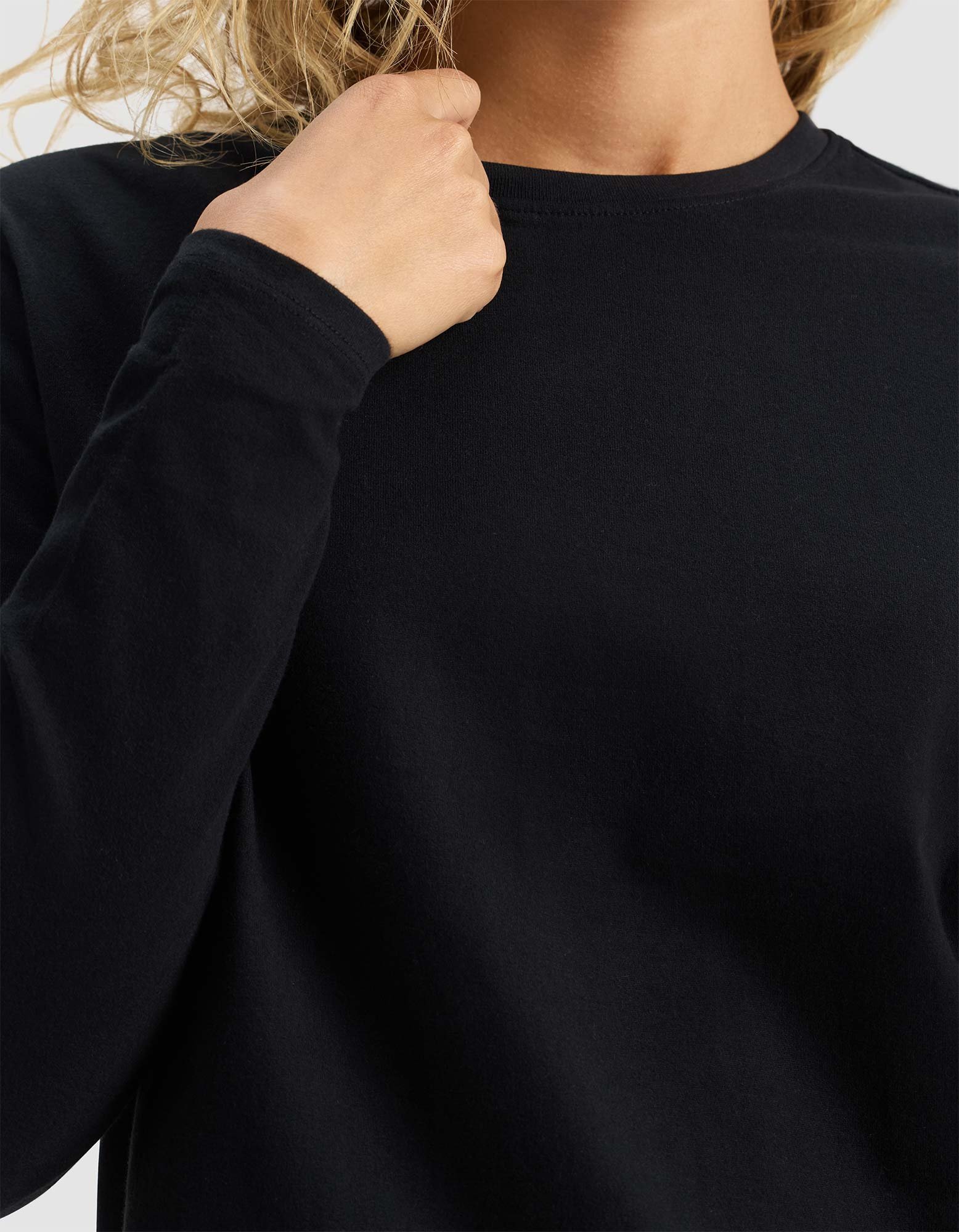 A woman wearing the Cozy Earth Women's All Day Long Sleeve Tee in black holds the neckline with one hand. Her face is hidden, and her light, wavy hair is partially visible.