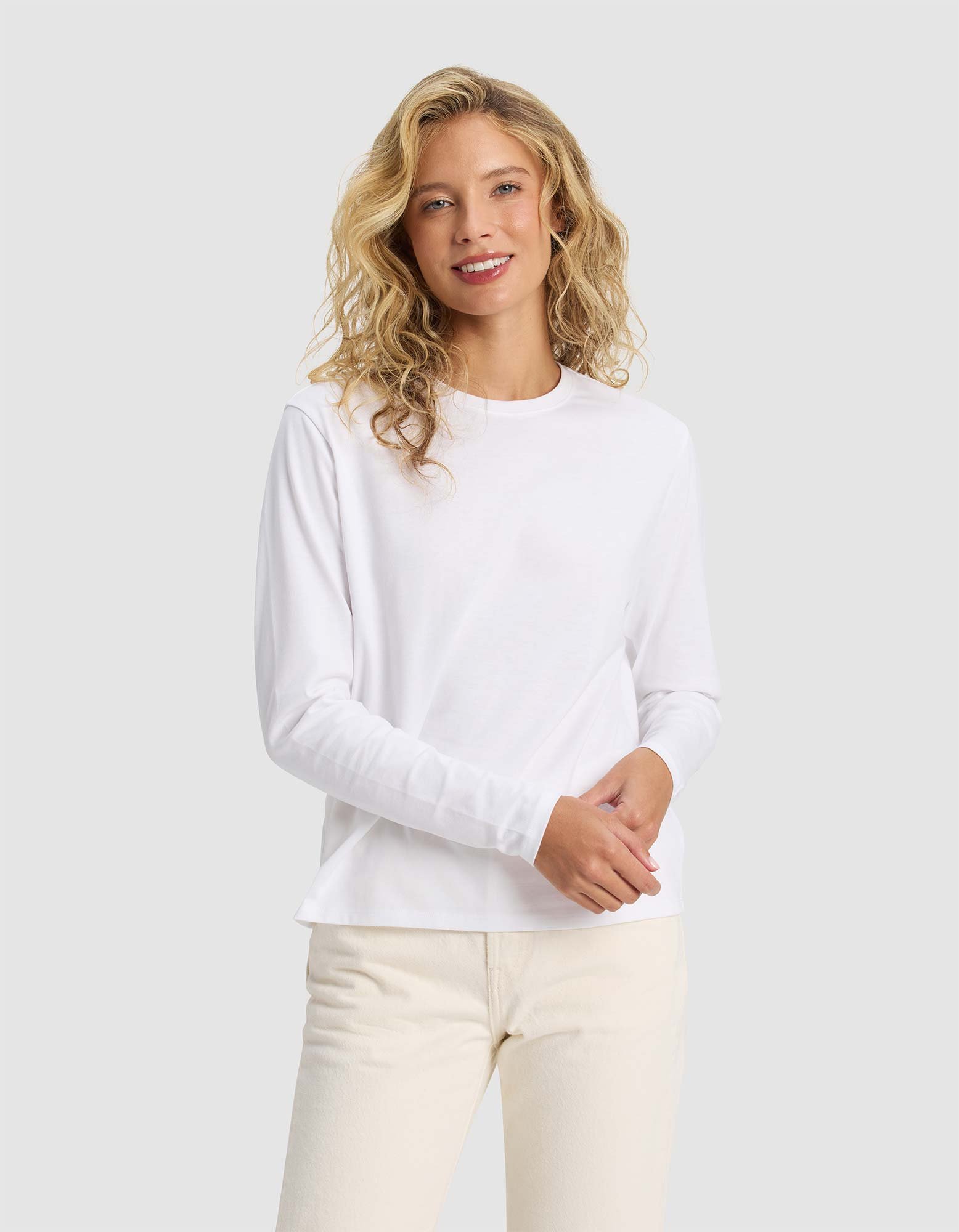 A woman with wavy blonde hair, dressed in Cozy Earth's Women's All Day Long Sleeve Tee and light-colored pants, stands against a light gray background, smiling softly at the camera.|Color:White