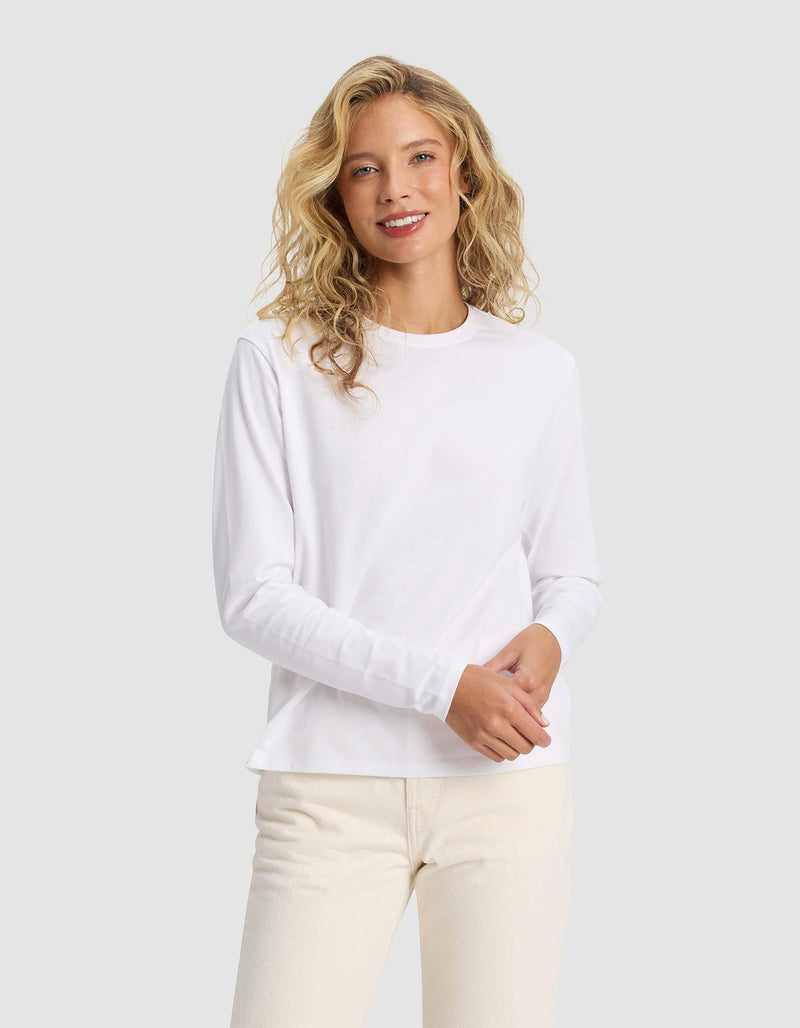A woman with wavy blonde hair, dressed in Cozy Earth's Women's All Day Long Sleeve Tee and light-colored pants, stands against a light gray background, smiling softly at the camera.