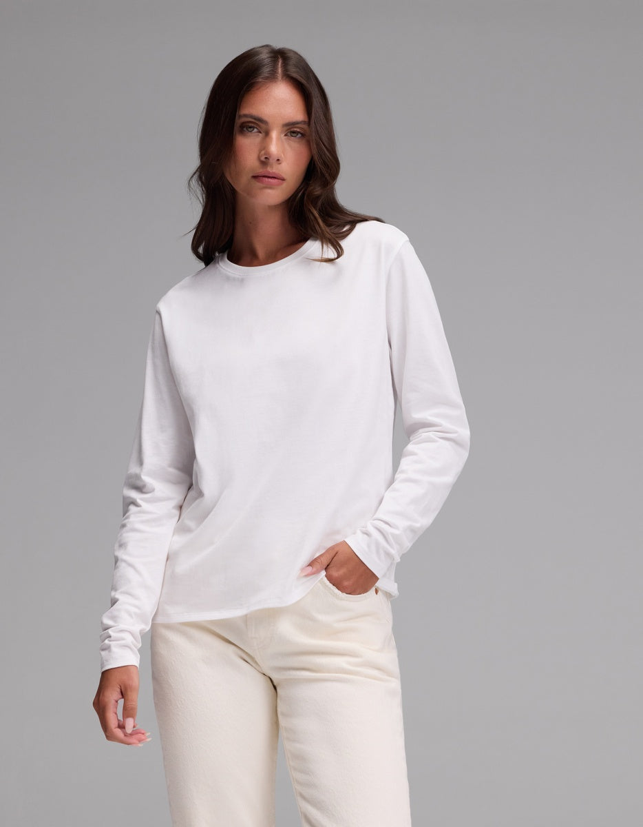 A person with long brown hair wears the Cozy Earth Women’s All Day Long Sleeve Tee in plain white and off-white pants, posing with one hand in their pocket against a plain gray background.