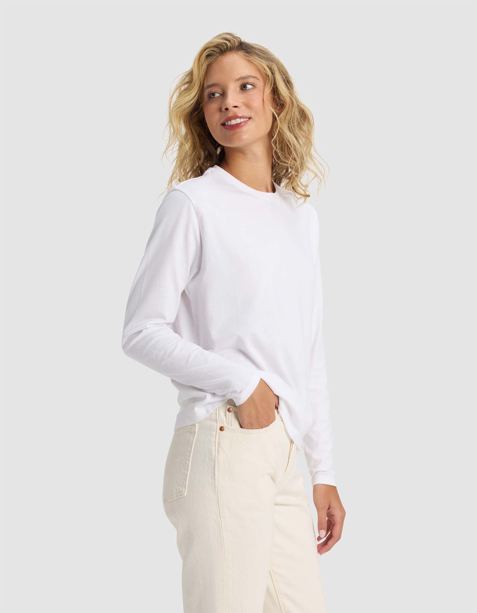 A woman with wavy blonde hair smiles as she models Cozy Earth's Women's All Day Long Sleeve Tee in white, paired with light beige pants and standing against a plain light gray background.|Color:White