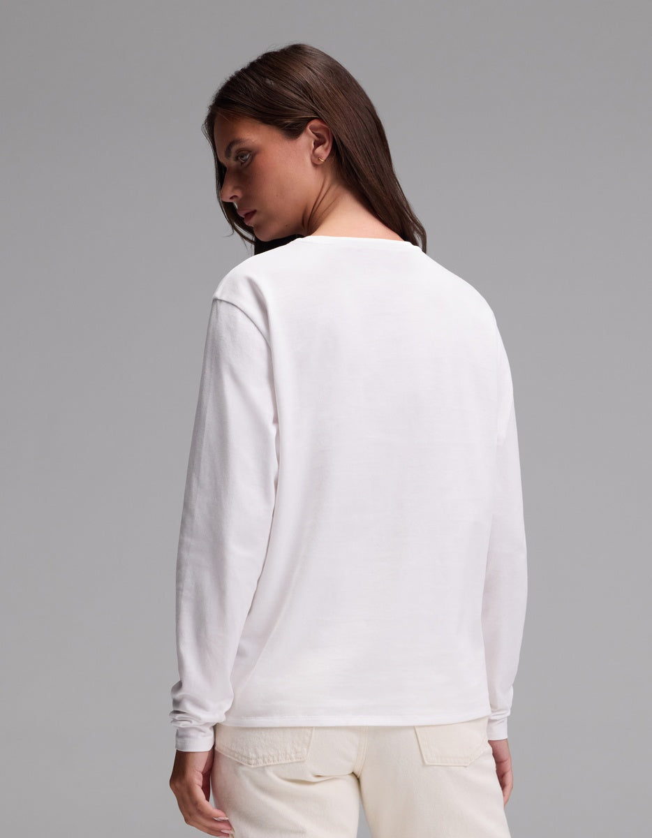 A woman wearing the Cozy Earth Women’s All Day Long Sleeve Tee in white and light-colored pants stands facing away from the camera, her long brown hair visible against a plain gray background.